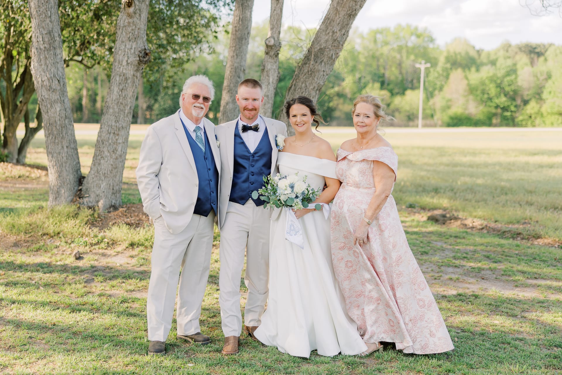 Bride and groom pose with two family members under large oak trees on the grounds of Scarborough House during a sunny wedding celebration.
