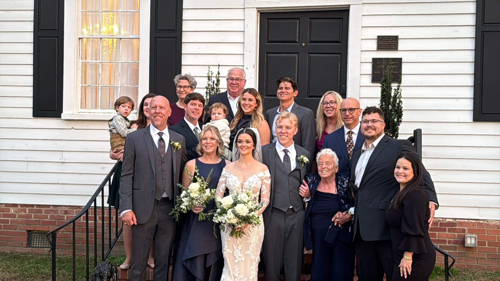 Bride and groom surrounded by smiling family members on the front steps of a white historic building with black shutters and door at Scarborough House wedding venue.