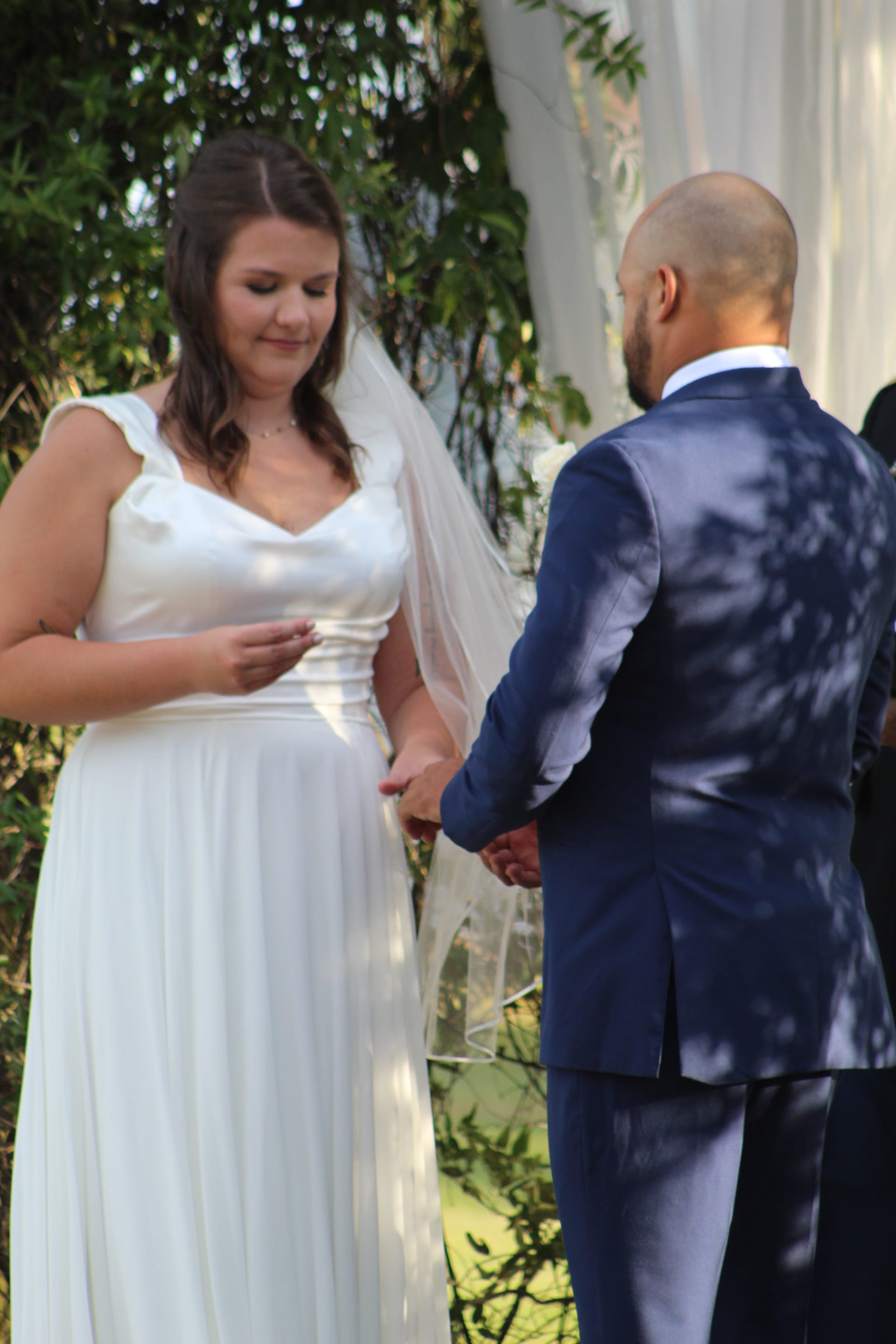Bride in a flowing white gown and veil smiles softly as she places a ring on the groom's hand during an outdoor wedding ceremony at Scarborough House.