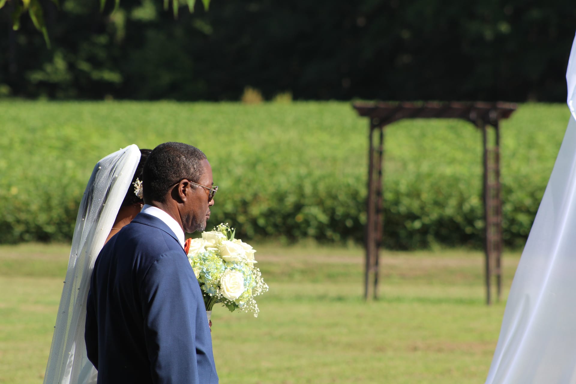 Groom in a navy suit walks beside his veiled bride holding a white and blue floral bouquet on the ceremony lawn at Scarborough House with a rustic wooden arch and green farmland fields in the background.