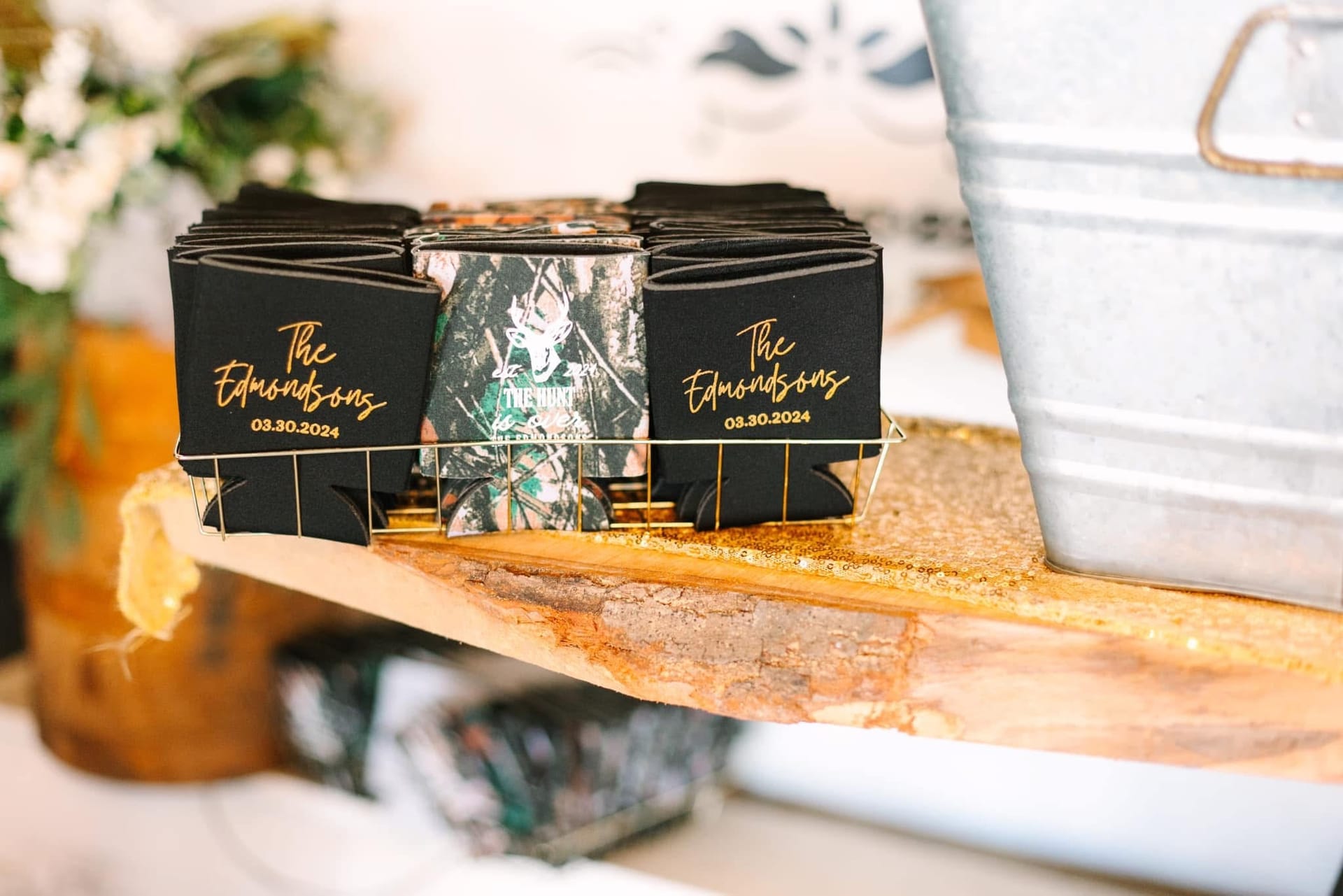 Custom black and camouflage wedding koozies printed with The Edmondsons and the date 03.30.2024 displayed in a wire rack on a live-edge wood board at Scarborough House.
