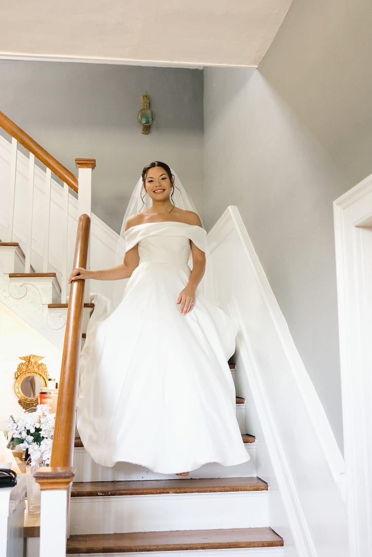 Radiant bride in an off-shoulder white ball gown and veil descends the grand wooden staircase of the 1821 Scarborough House manor, smiling warmly at the camera.