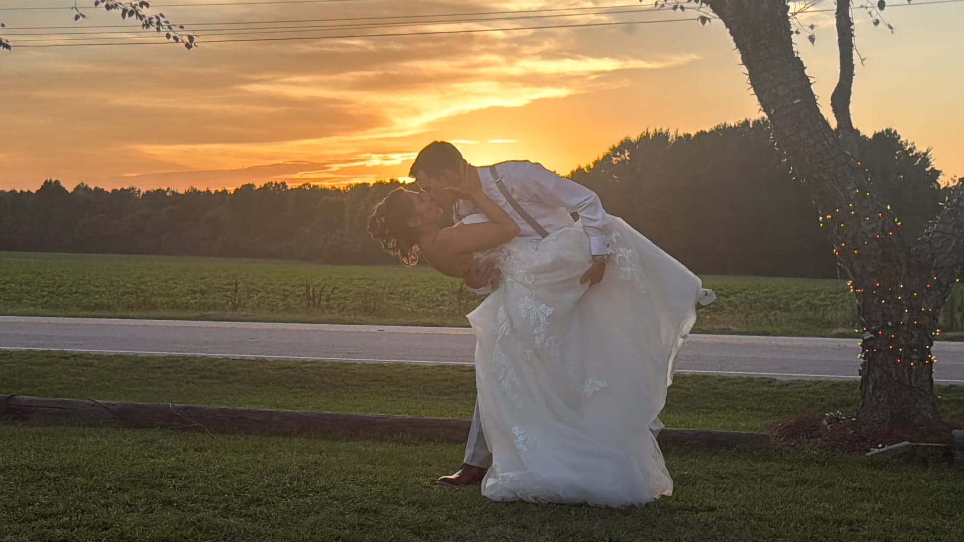Bride and groom share a romantic dip kiss silhouetted against a vivid orange and gold sunset sky with open farmland fields and tree line in the background at Scarborough House.