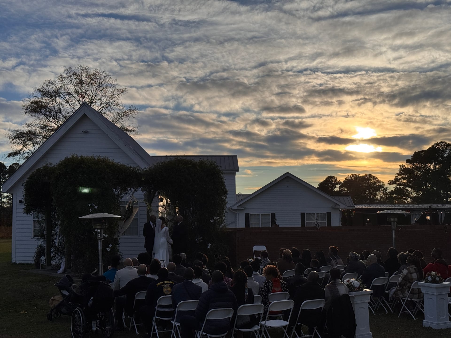 Outdoor wedding ceremony at dusk on the Scarborough House ceremony lawn with guests seated in white folding chairs facing the couple at a vine-covered archway beneath a dramatic golden sunset sky.
