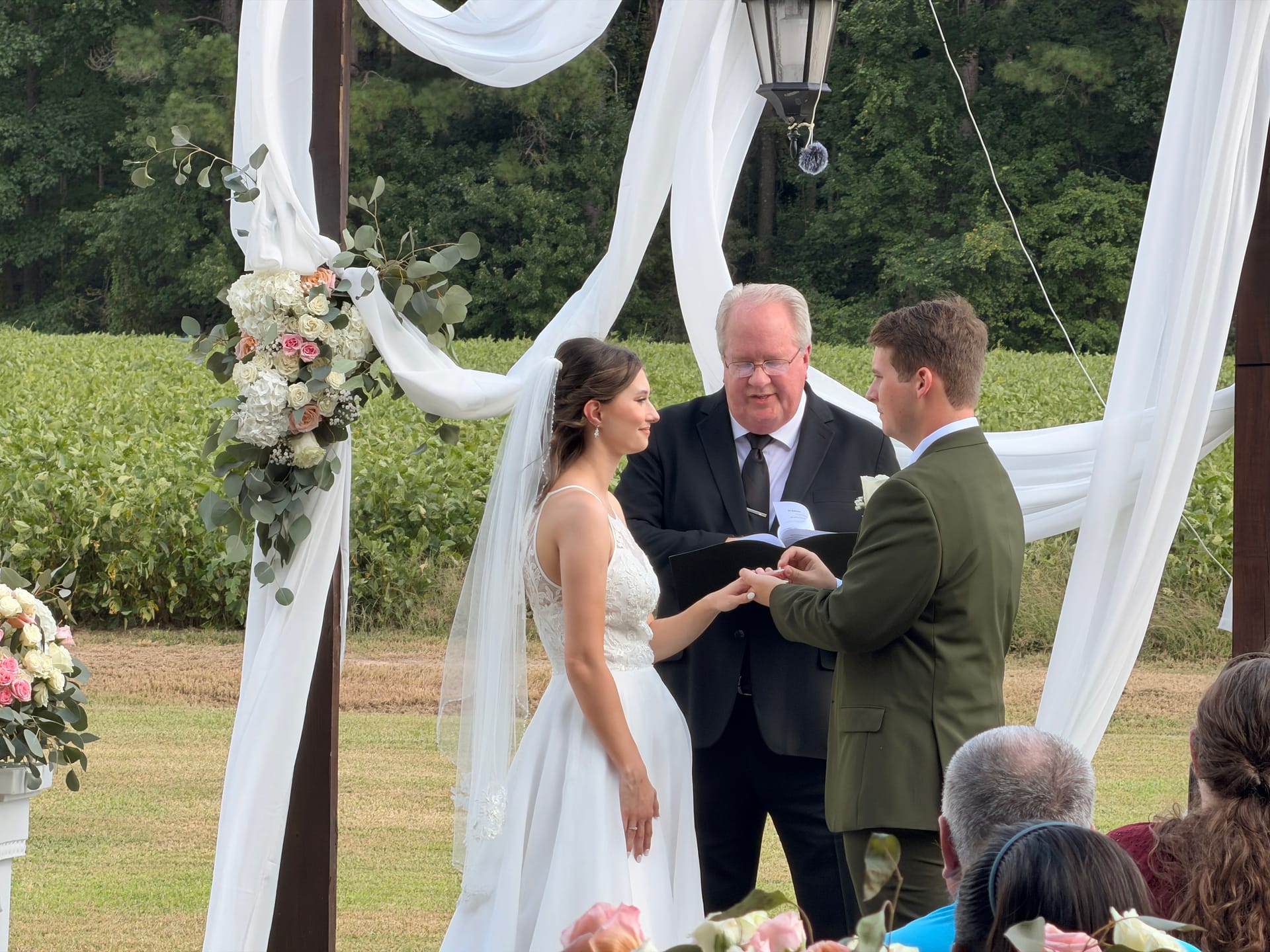Bride and groom exchange rings beneath a white-draped floral arch on the ceremony lawn at Scarborough House, with lush green farmland fields in the background.