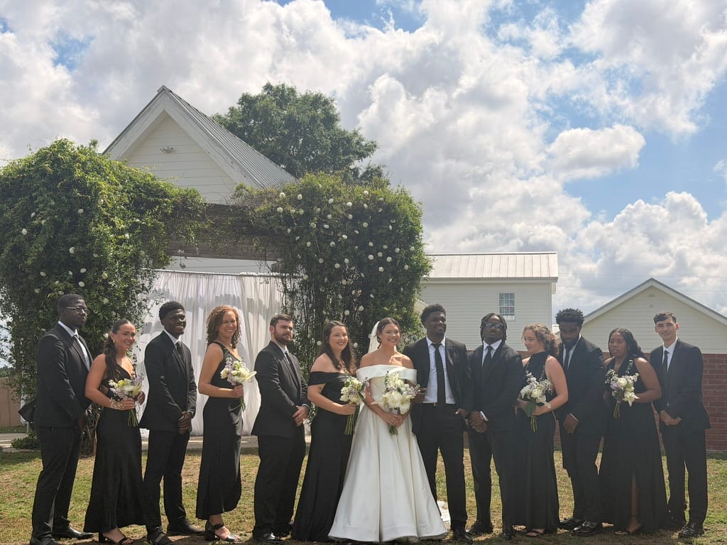 Bride and groom surrounded by their wedding party dressed in black formal attire posing in front of a vine-covered floral arch at Scarborough House ceremony lawn.
