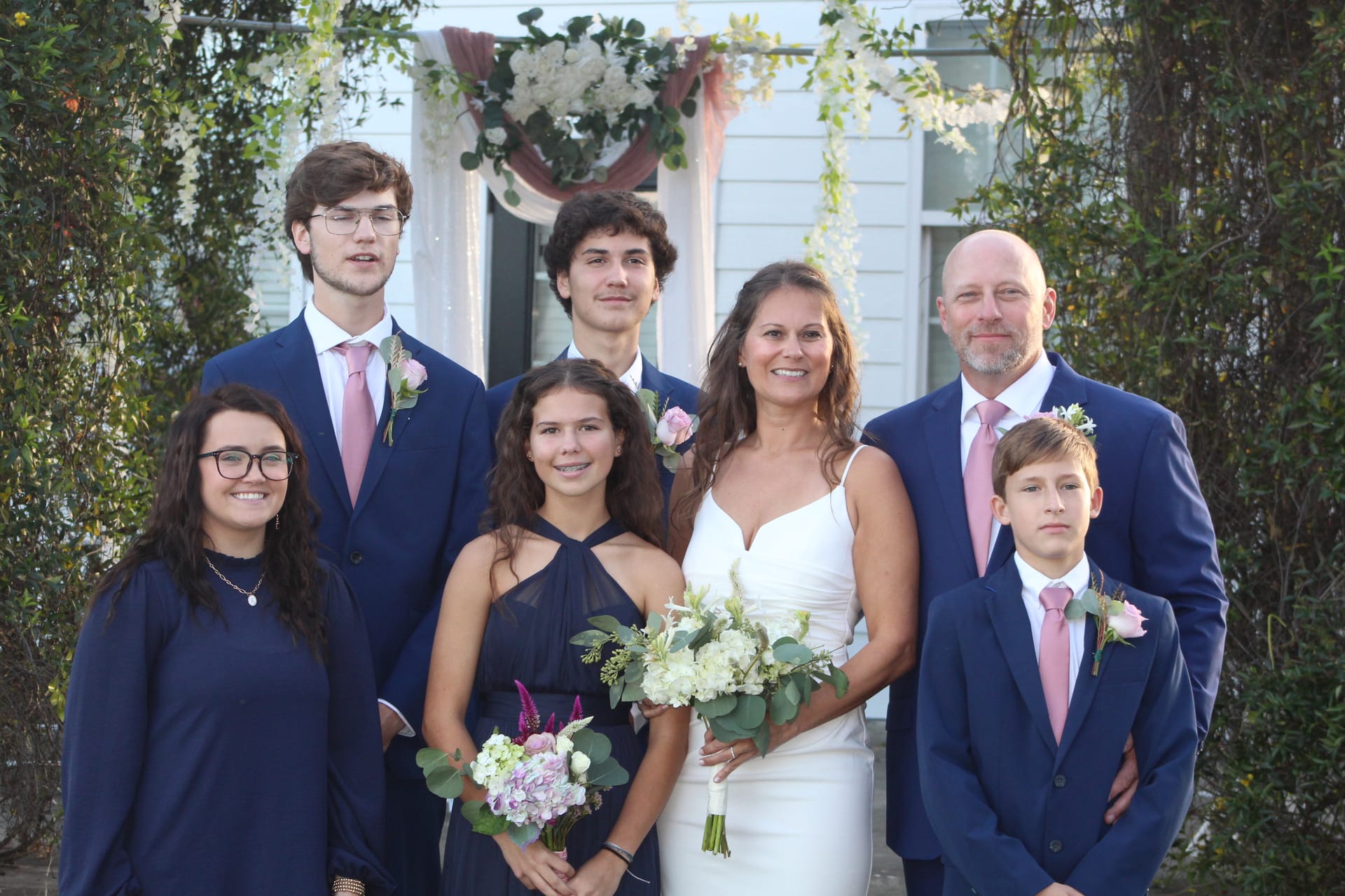 Bride in white gown and groom in navy suit pose with five children in coordinating navy and pink attire beneath a floral arch at the entrance of a historic estate wedding venue.