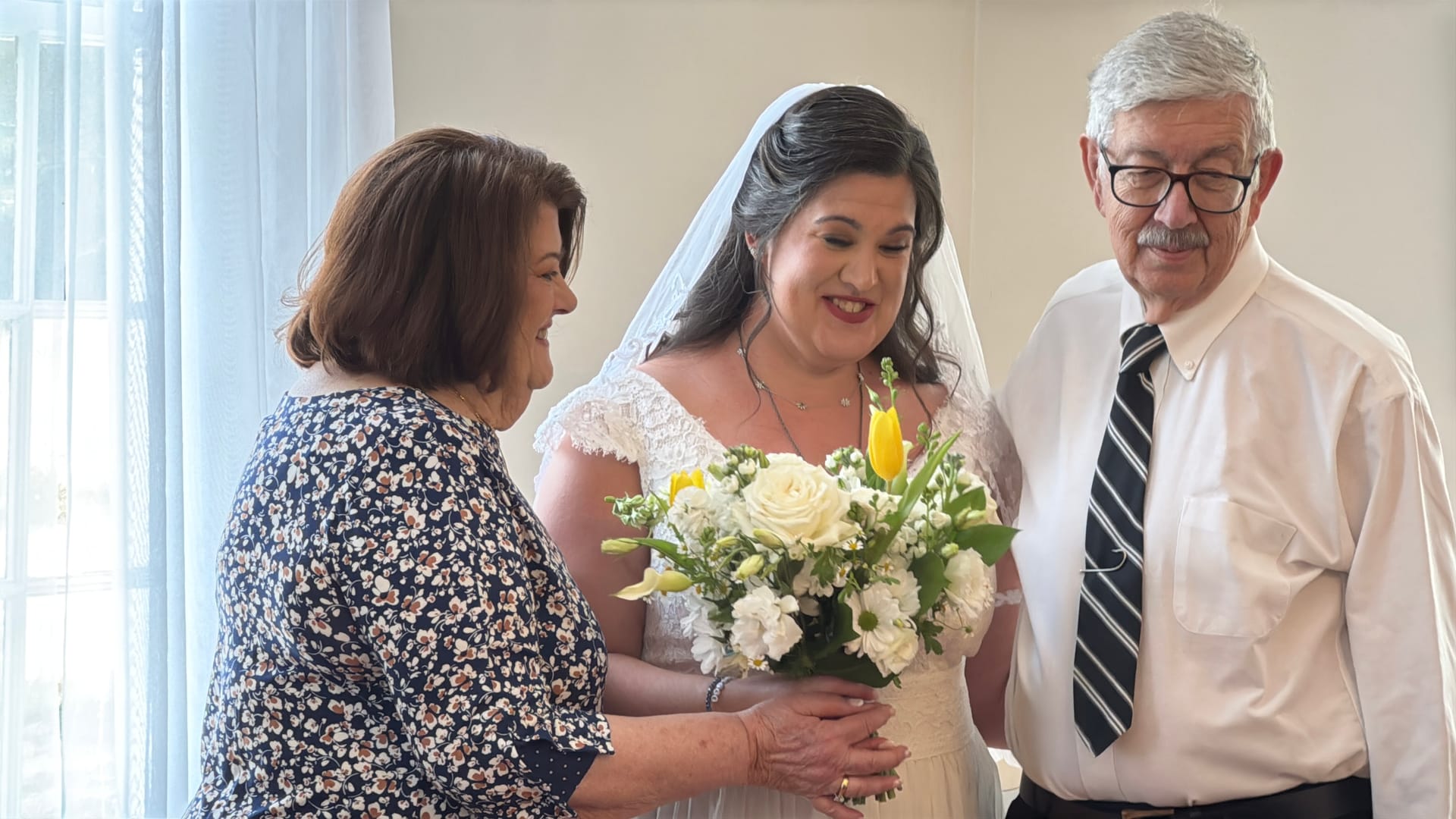 A bride in a white lace gown and veil shares a joyful moment with an older woman and man as they admire her white and yellow floral bouquet indoors before the ceremony.