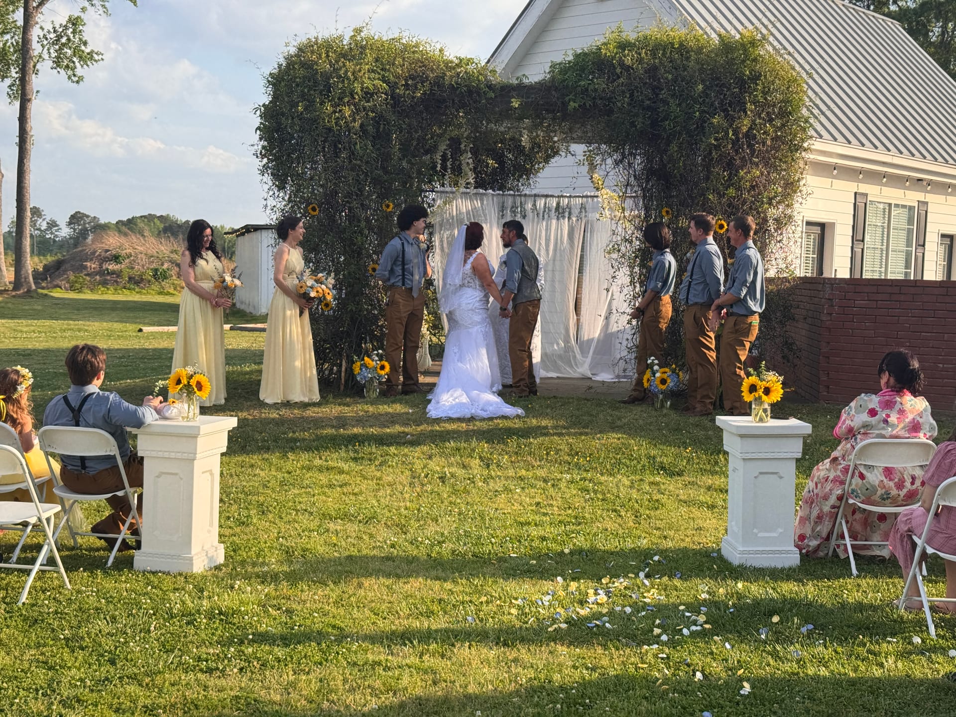 Bride and groom exchange vows beneath a lush greenery arch adorned with white draping and sunflowers on the ceremony lawn at Scarborough House, flanked by bridesmaids in yellow gowns and groomsmen in denim and tan.