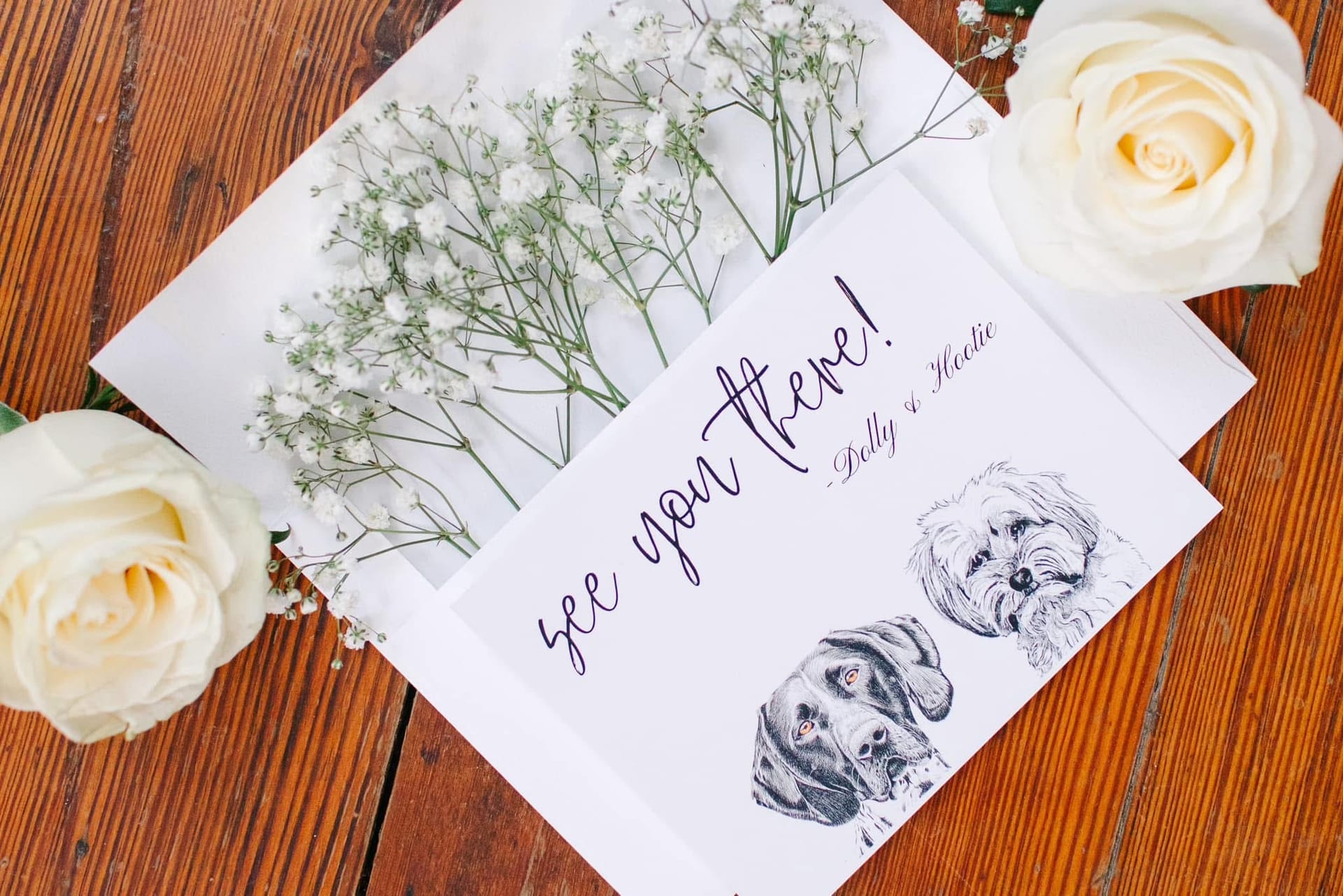 Elegant wedding flat-lay featuring a calligraphy see you there card signed by dogs Dolly and Hoodie with custom pet portraits, white roses, and baby's breath arranged on a rustic wood surface.