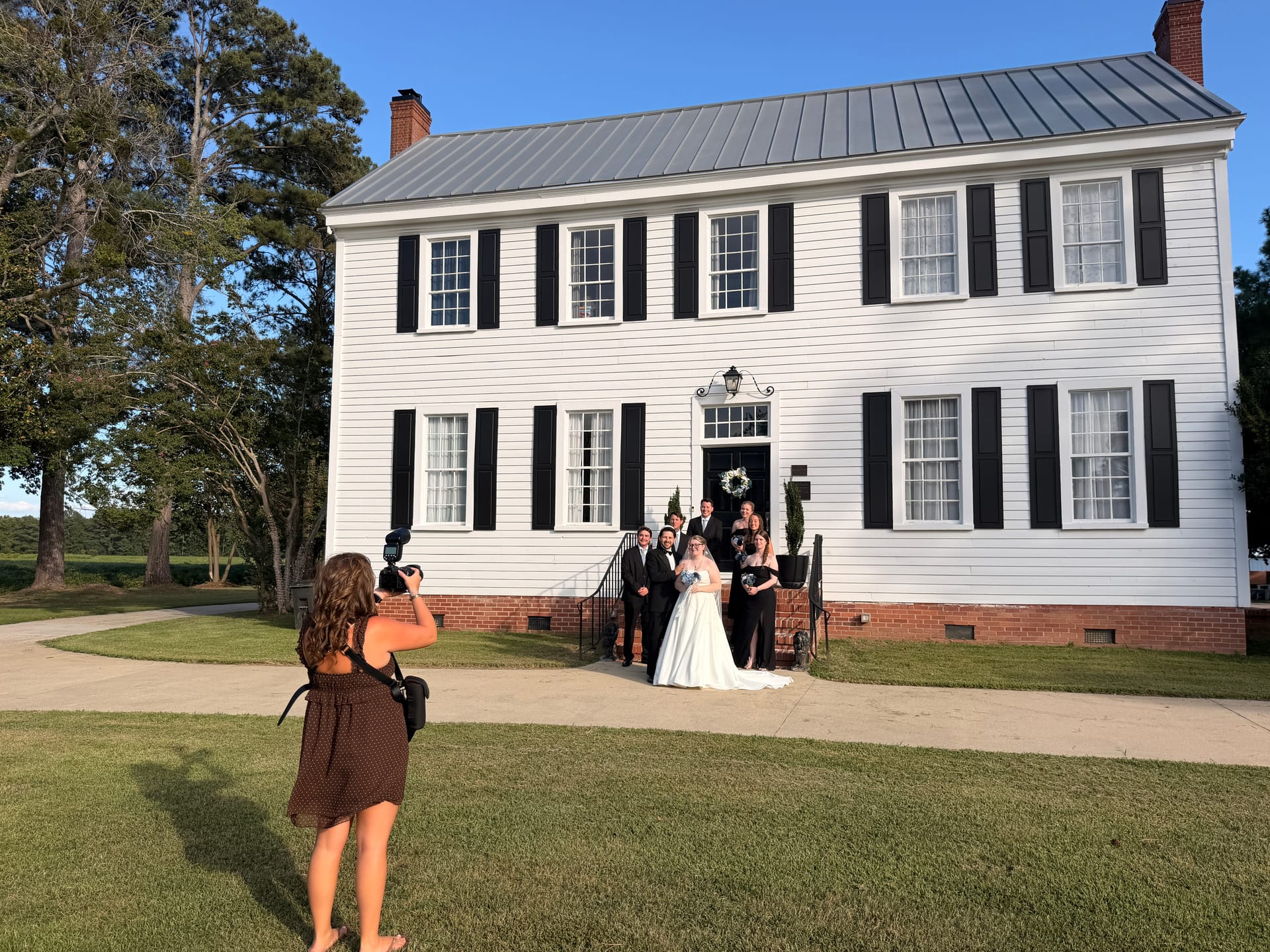 A wedding photographer captures the bride, groom, and wedding party posing on the front steps of the 1821 Scarborough House manor under warm golden-hour light.