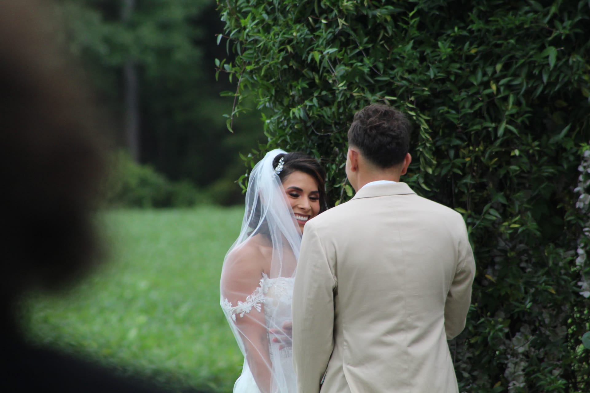 A beaming bride in a lace off-shoulder gown and veil shares a joyful moment with her groom during an outdoor garden ceremony at Scarborough House, surrounded by lush green hedges.