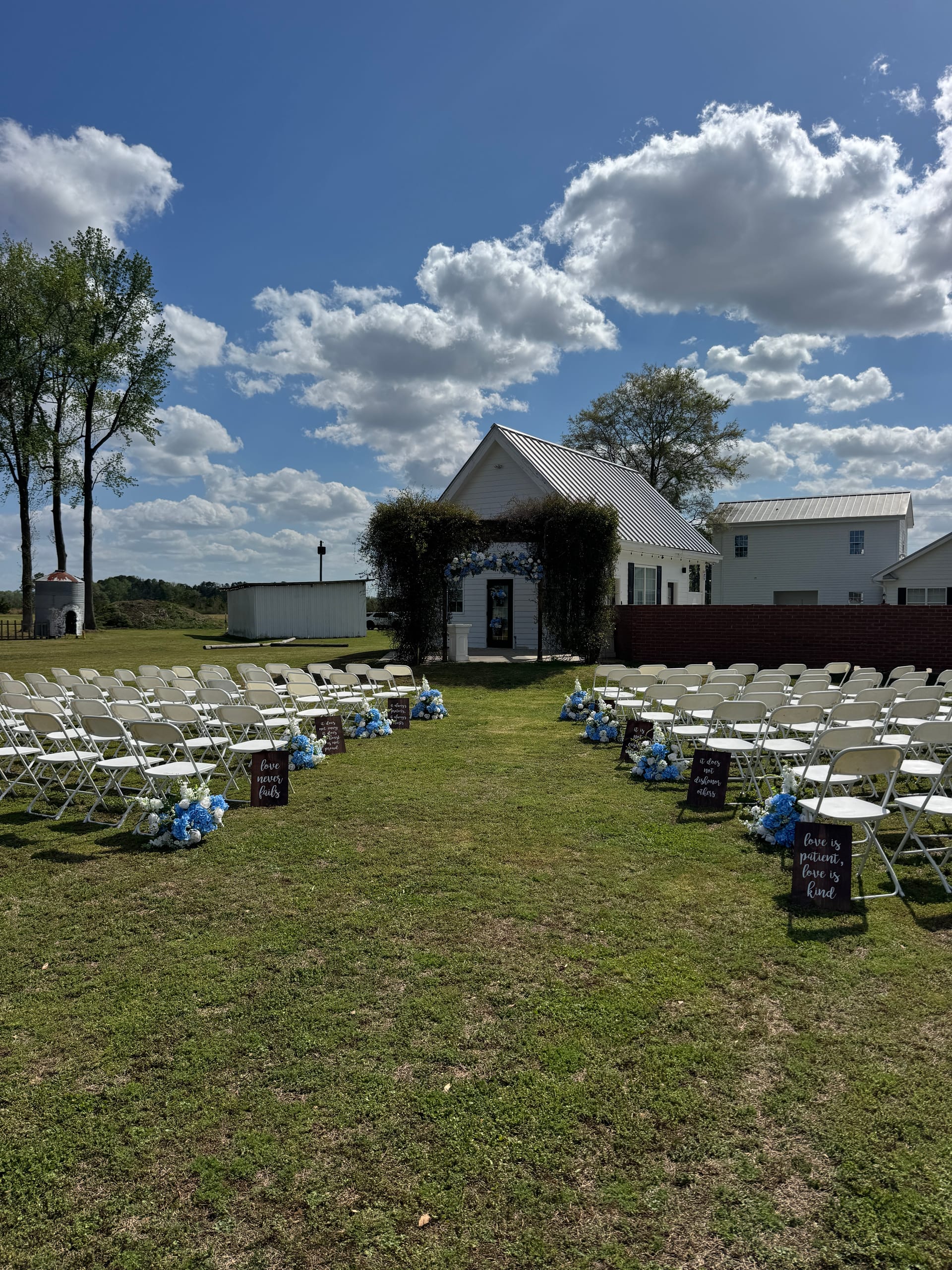 Rows of white folding chairs lined with blue hydrangea floral arrangements and wooden signs lead down a grass aisle toward a white chapel adorned with a floral arch at Scarborough House wedding venue.