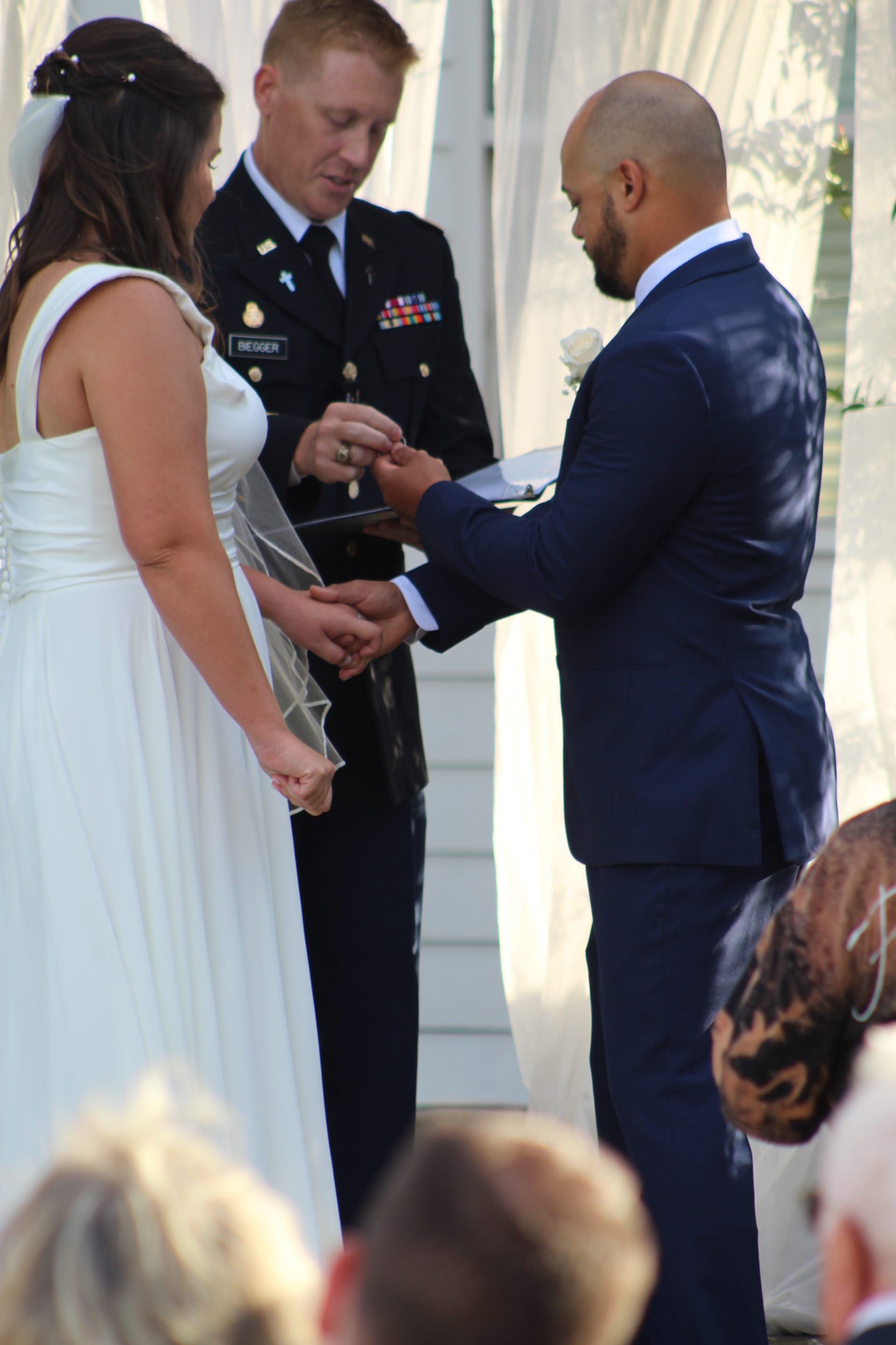 Groom places a wedding ring on the bride's finger during an outdoor ceremony officiated by a military officer at a historic estate venue.