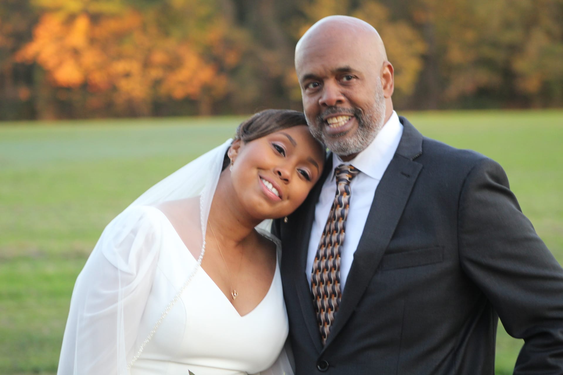 Bride in white gown and veil rests her head on a suited gentleman's shoulder as both smile warmly against a golden autumn landscape at Scarborough House.