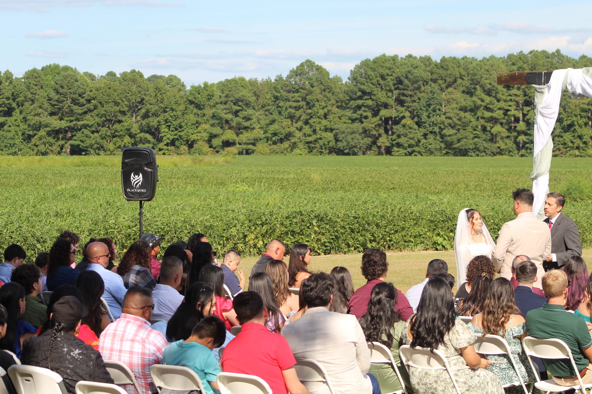 Guests seated in white folding chairs watch an outdoor wedding ceremony at a rustic farmland venue with lush green fields and wooded tree line in the background.