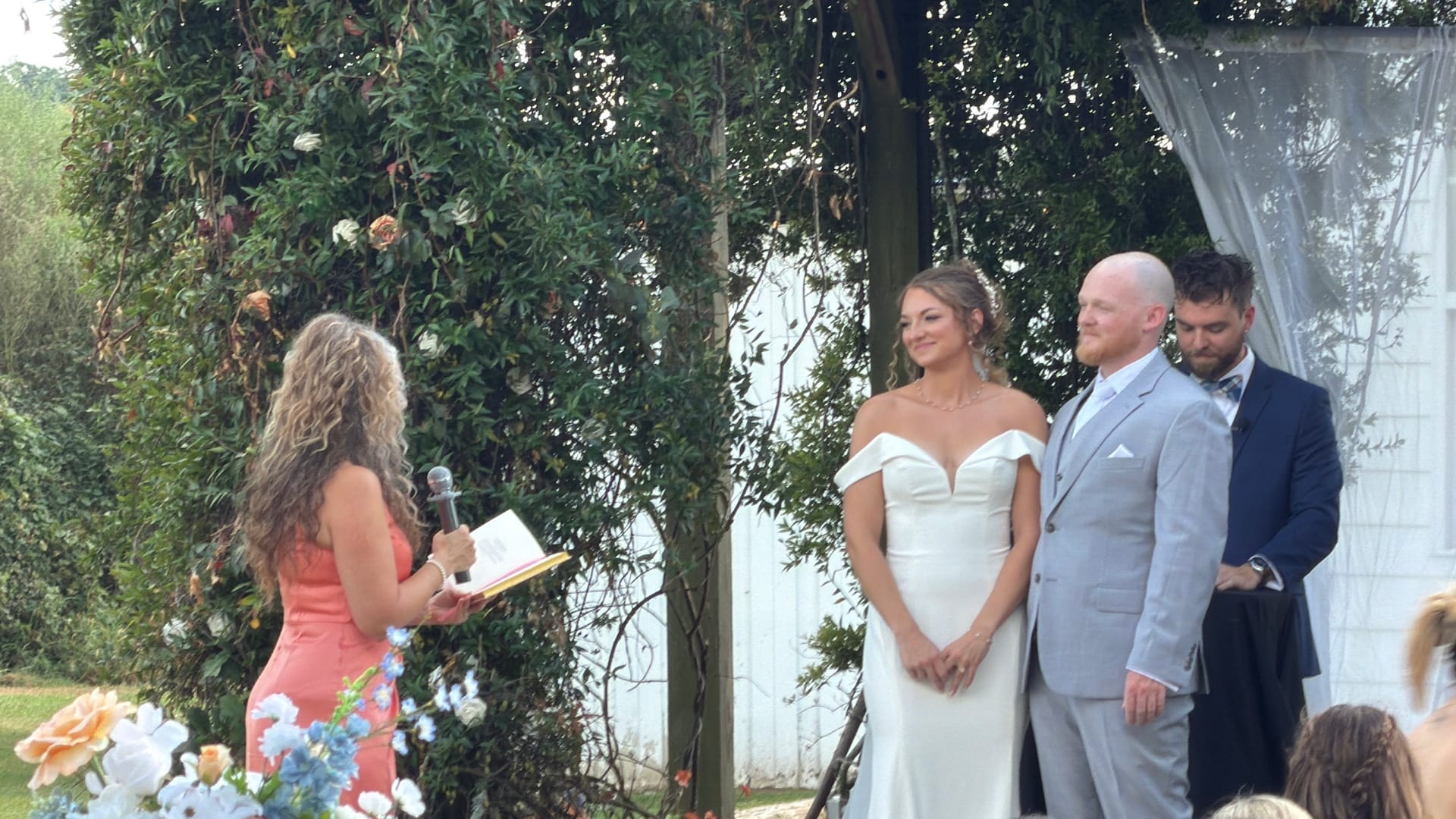 Smiling bride in an off-shoulder white gown and groom in a light grey suit stand at the altar during their outdoor garden wedding ceremony at Scarborough House while the officiant reads from her notes.