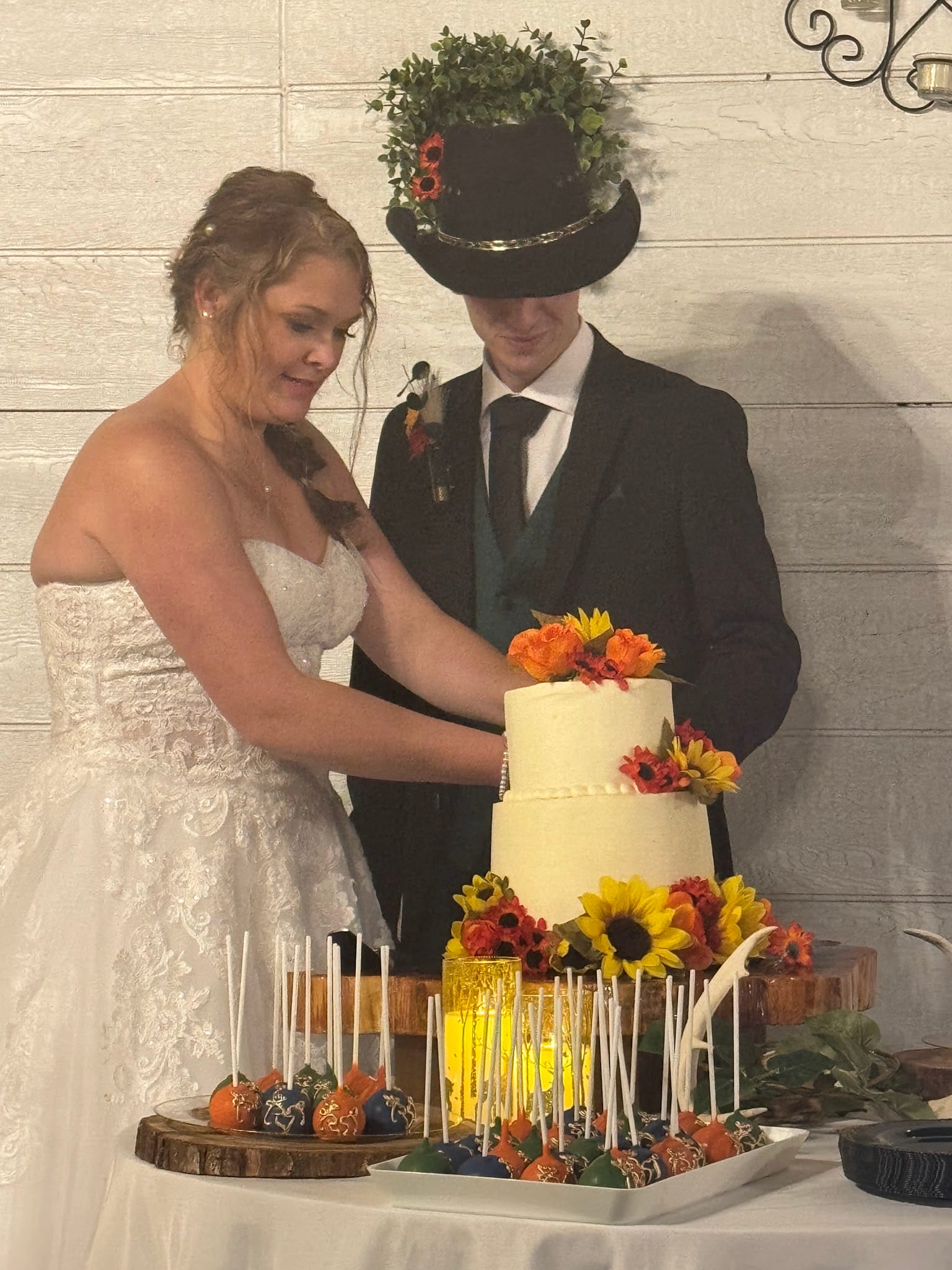 Bride in a white lace gown and groom in a black suit with a floral-adorned top hat cut their two-tier white wedding cake decorated with sunflowers and autumn blooms at Scarborough House.