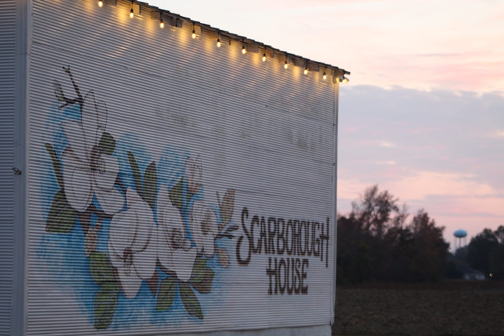 Scarborough House wedding venue exterior at dusk featuring a colorful mural and warm string lights along the roofline against a soft pink and purple sunset sky.