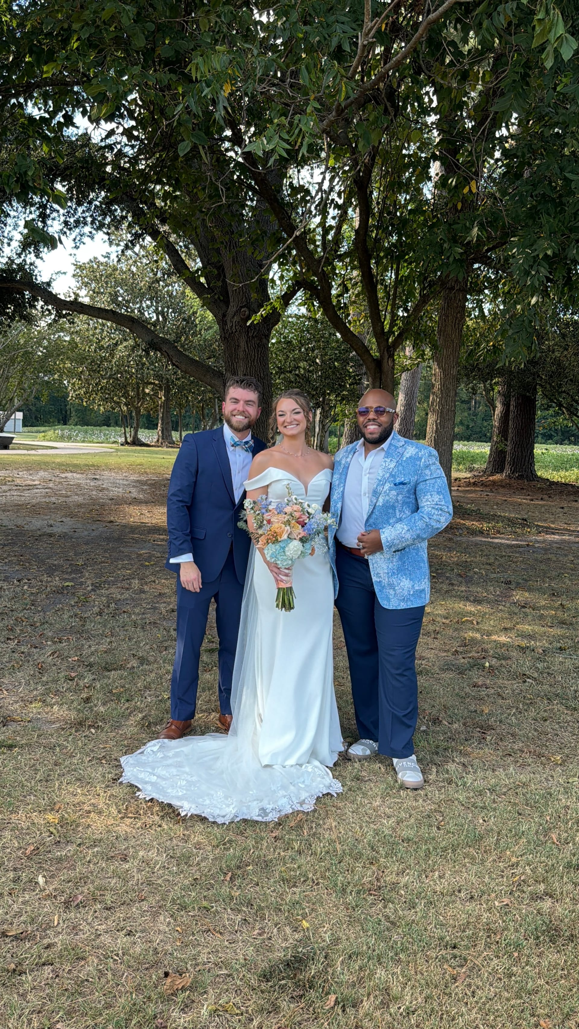 Bride in an off-shoulder white gown holding a pastel bouquet stands between two guests in blue suits beneath the oak trees at Scarborough House estate.