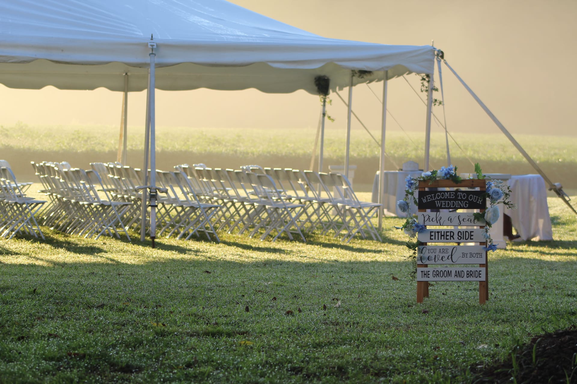 A rustic wooden wedding welcome sign adorned with blue flowers reads Pick a Seat Either Side You Are Loved by Both the Groom and Bride, set before rows of white folding chairs under a large white tent on the ceremony lawn in warm golden morning light.