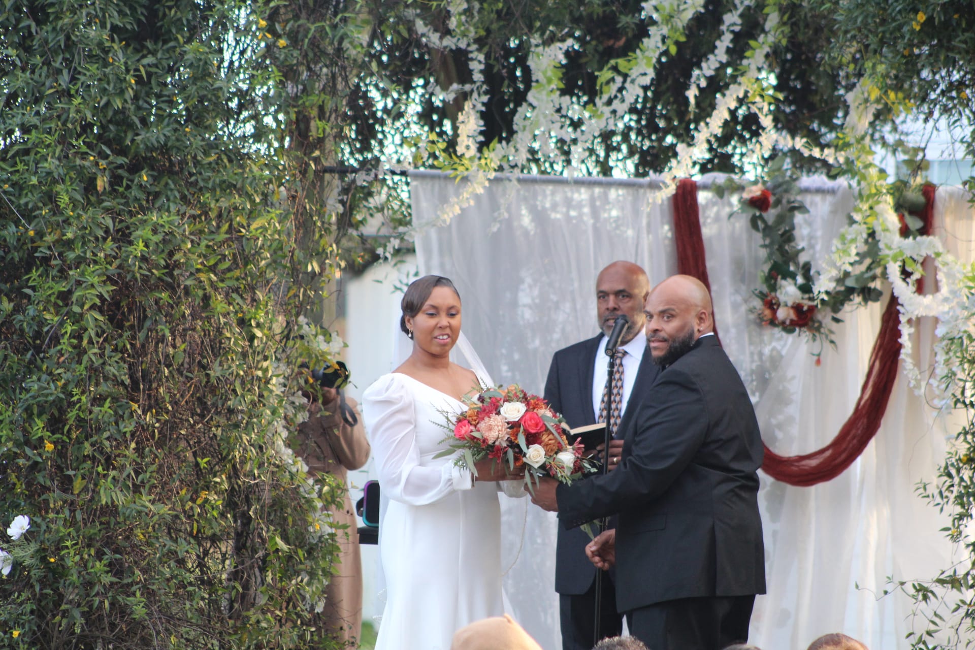 Bride in white gown holding a colorful bouquet exchanges vows with groom in black suit before an officiant at a flower-adorned outdoor wedding arch at Scarborough House.