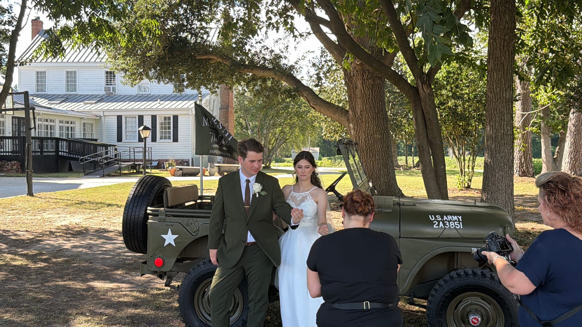 Bride and groom pose together in front of a vintage U.S. Army jeep beneath oak trees on the historic grounds of Scarborough House wedding venue while a photographer captures the moment.