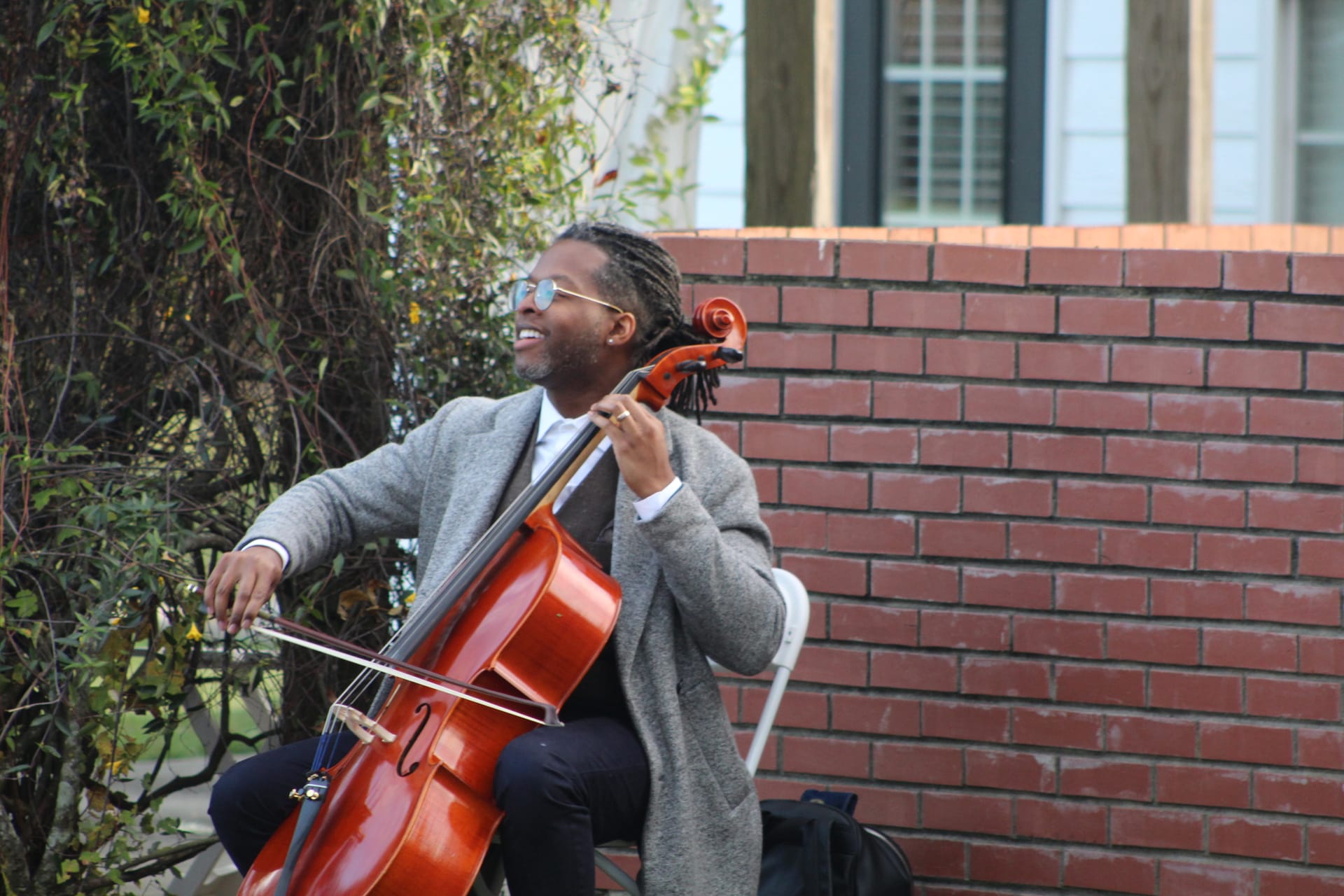 A smiling cellist in a gray coat plays cello outdoors against a red brick wall at a historic estate wedding venue.