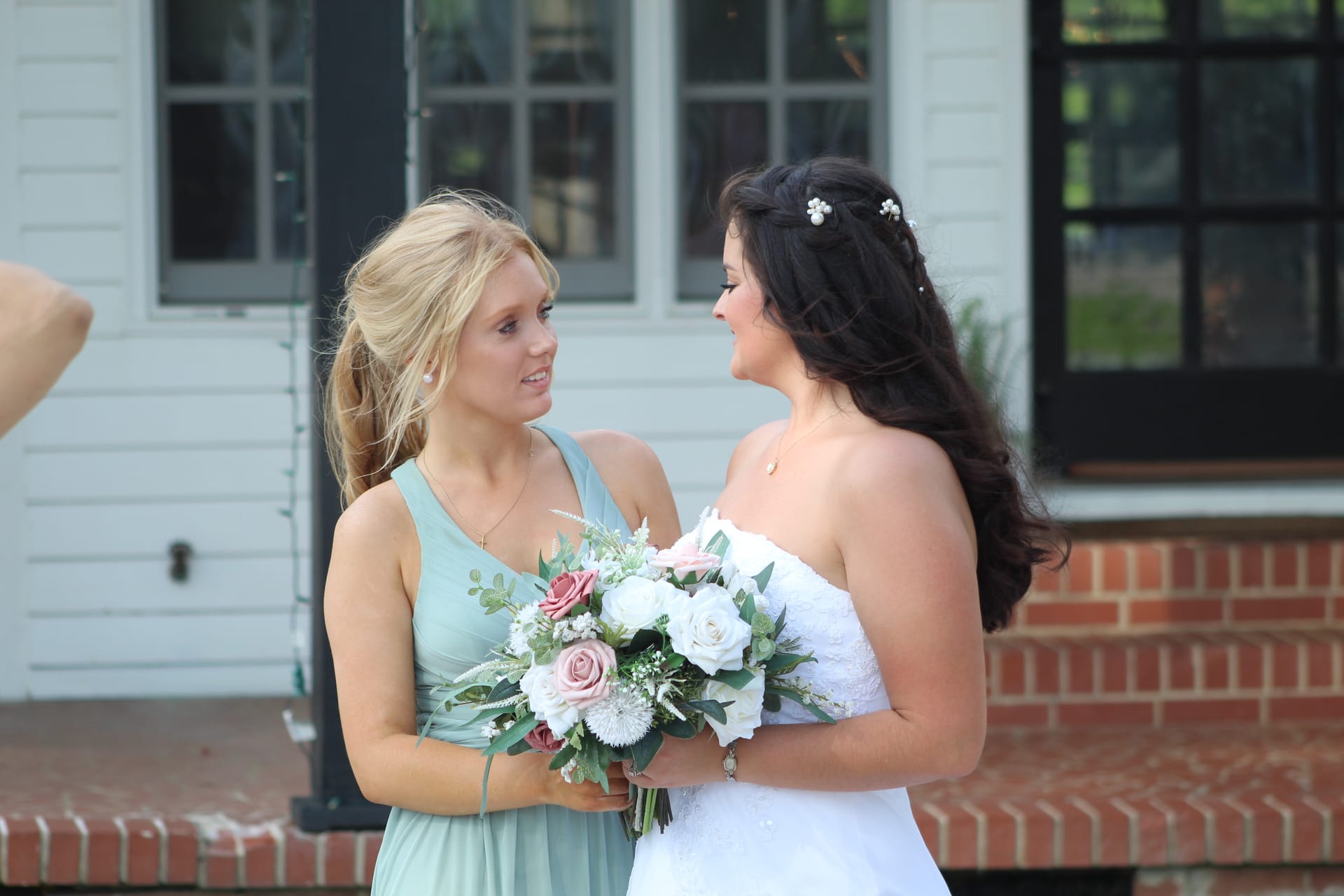 Bride in white lace gown holding a pink and white rose bouquet shares a warm moment with her bridesmaid in a sage green dress outside a historic white manor house.