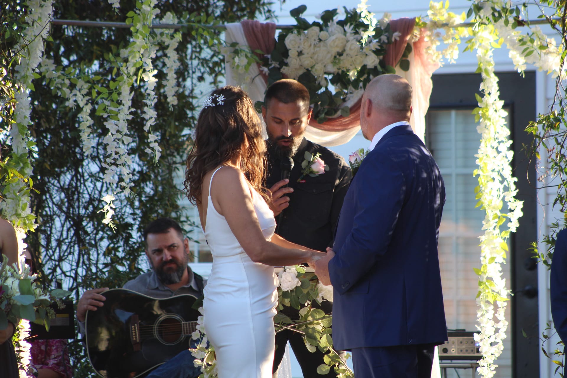Bride and groom exchange vows beneath a lush floral arch adorned with white wisteria and greenery at Scarborough House while a guitarist plays in the background.