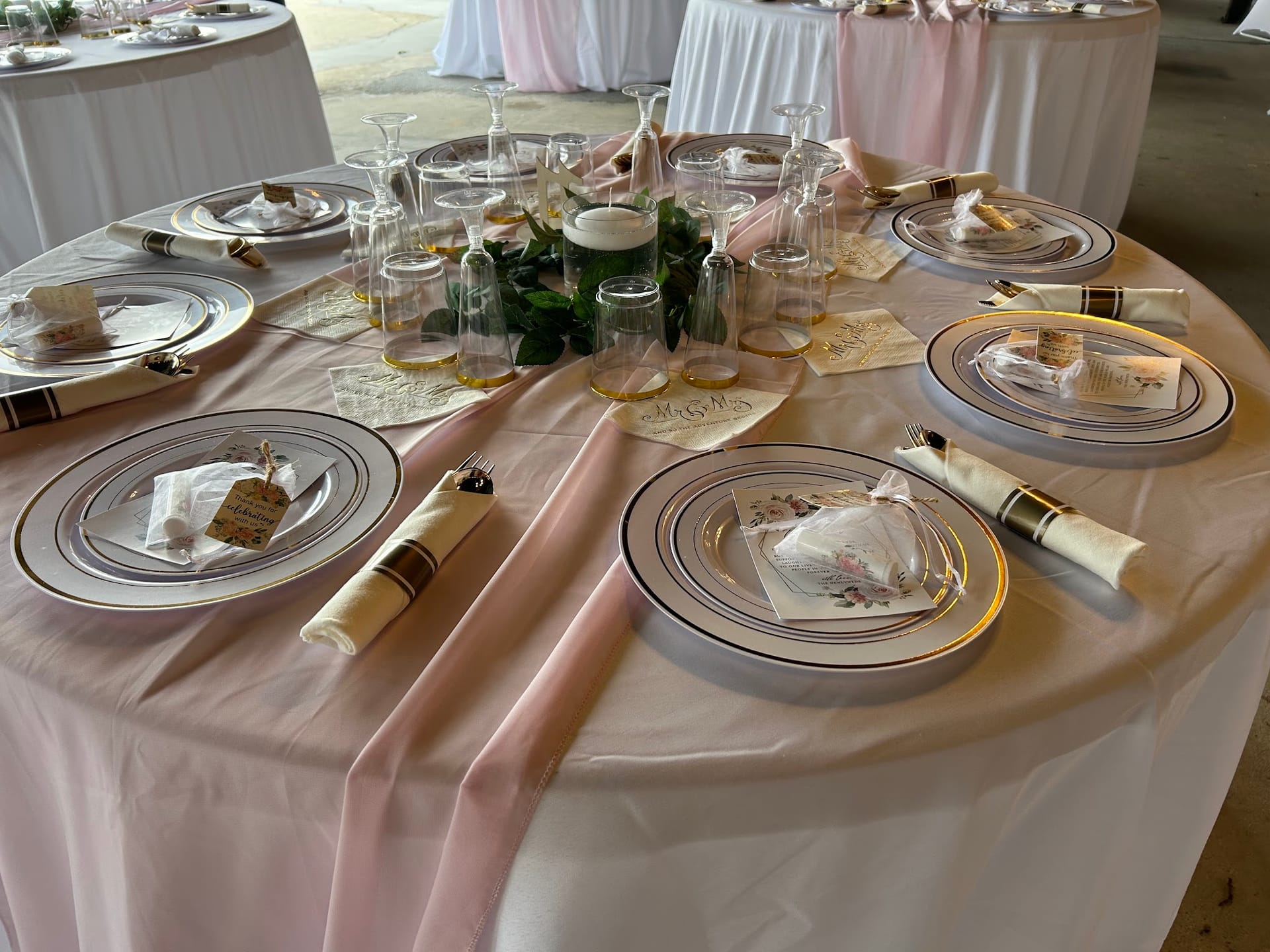 Elegantly set round reception table with blush pink linens, gold-rimmed white charger plates, rolled napkins with place cards, and a greenery and candle centerpiece surrounded by clear glassware.