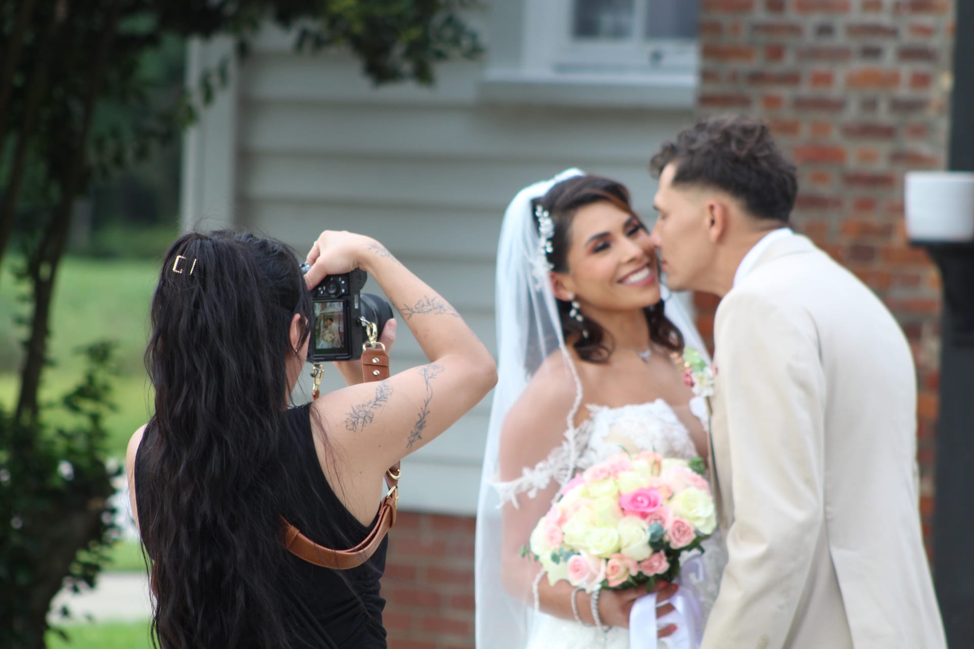 A wedding photographer captures a smiling bride holding a pink and white rose bouquet as the groom kisses her cheek outside a historic brick and clapboard estate.