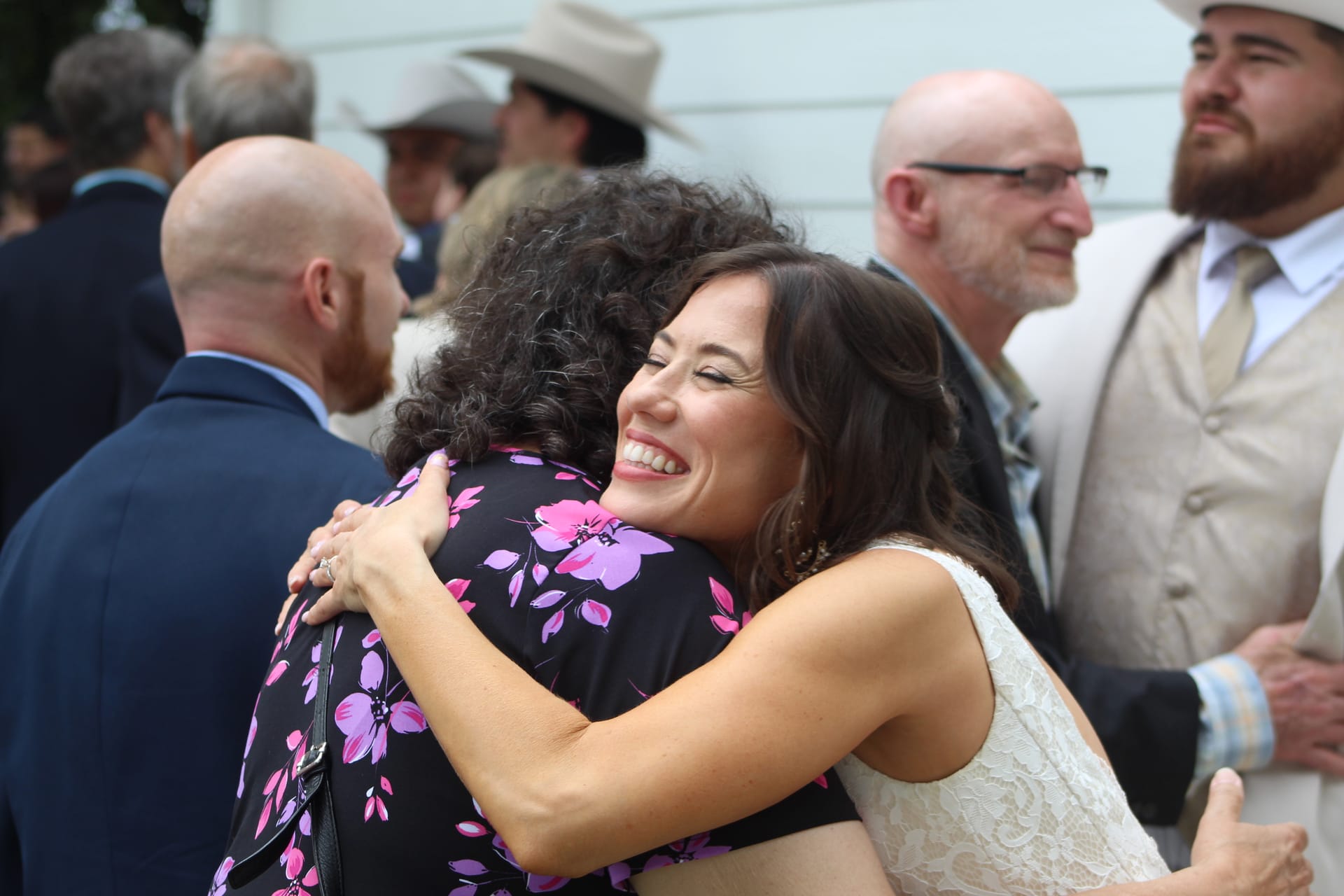 Bride in white lace dress laughs warmly while hugging a guest in a floral dress during an outdoor wedding reception at a historic estate venue.