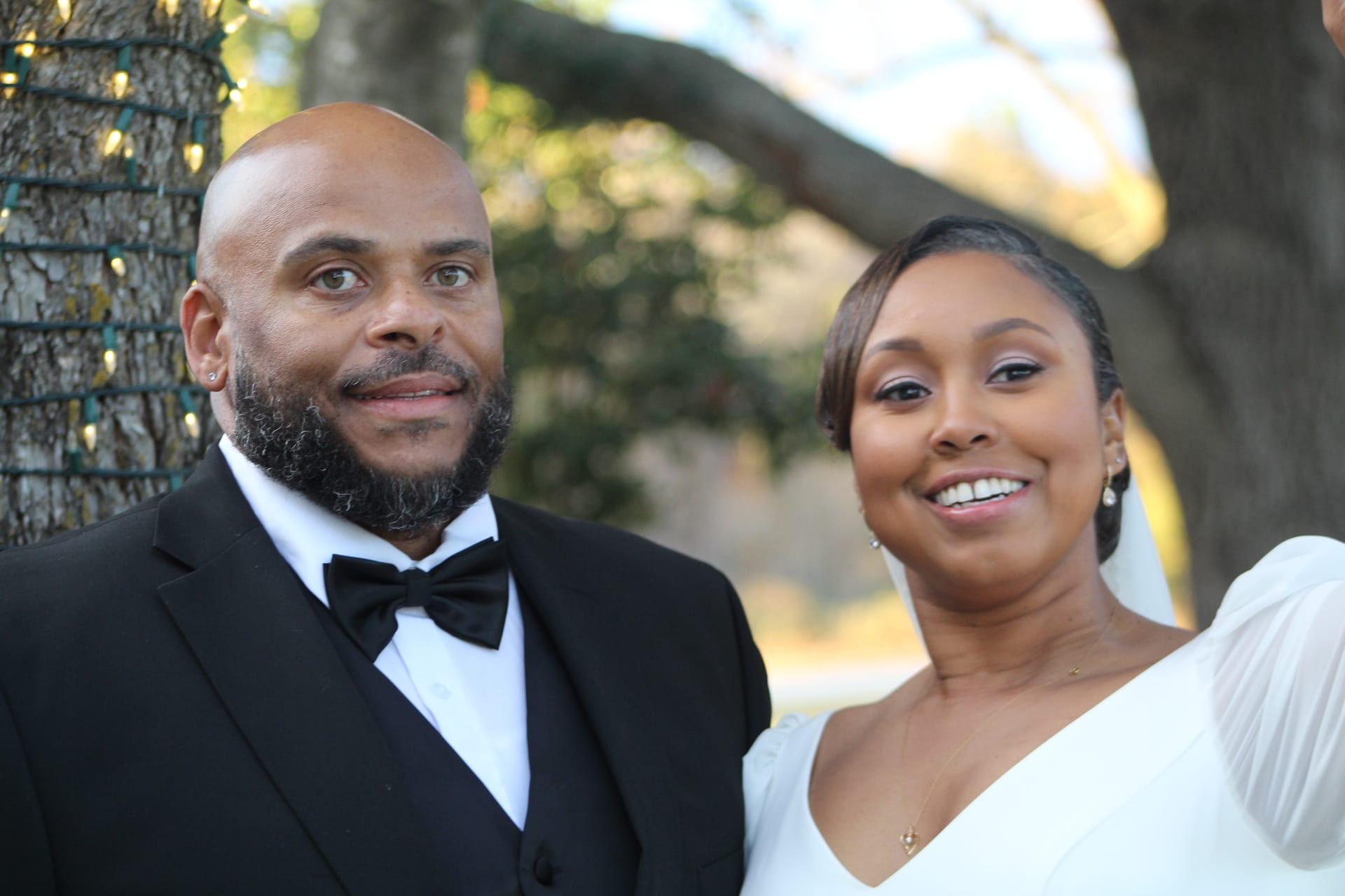 Bride in a white gown and groom in a black tuxedo with bow tie smile together in front of a grand oak tree at Scarborough House wedding venue.