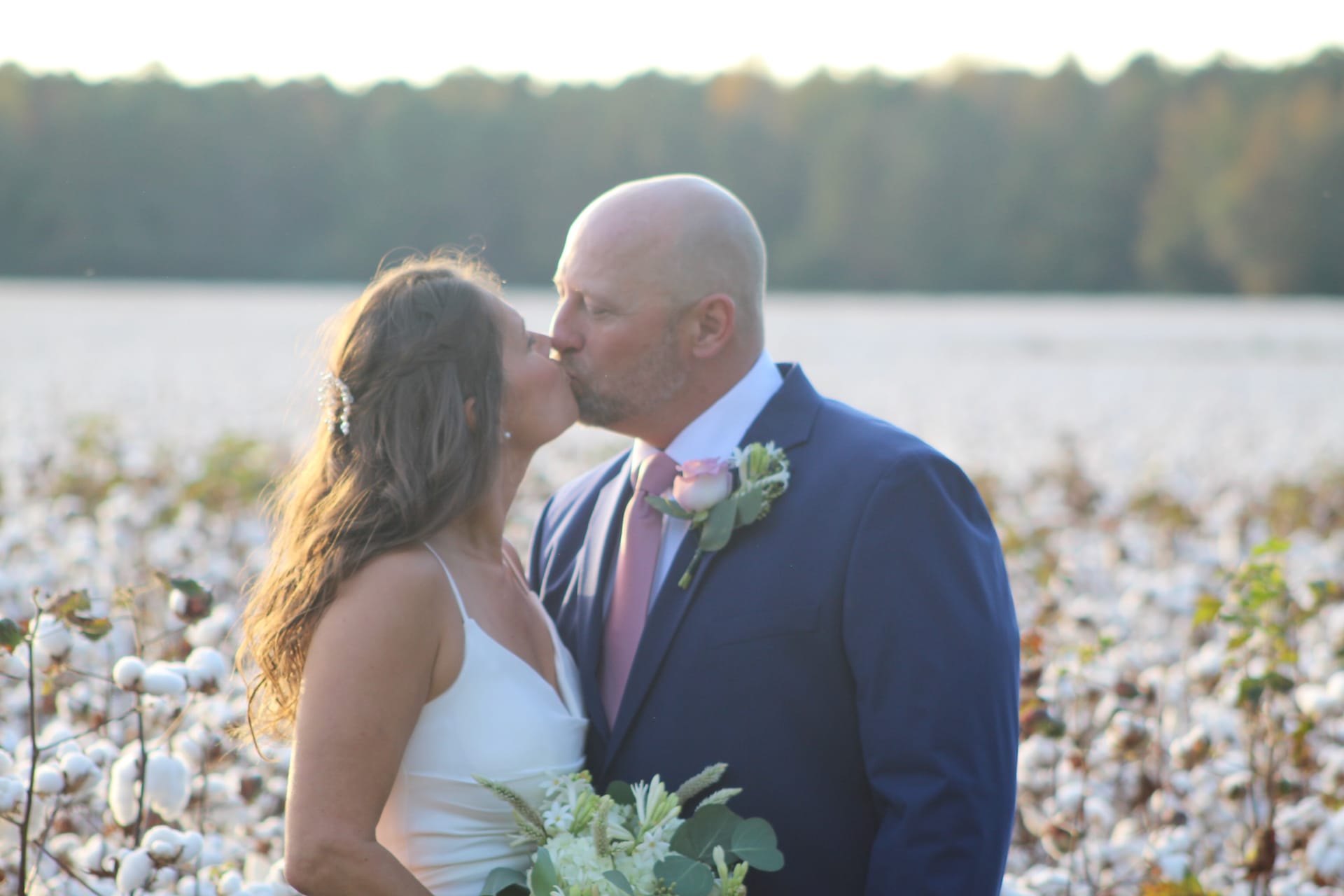 Bride and groom share a kiss among cotton field rows at golden hour with a tree line glowing softly in the background at Scarborough House.