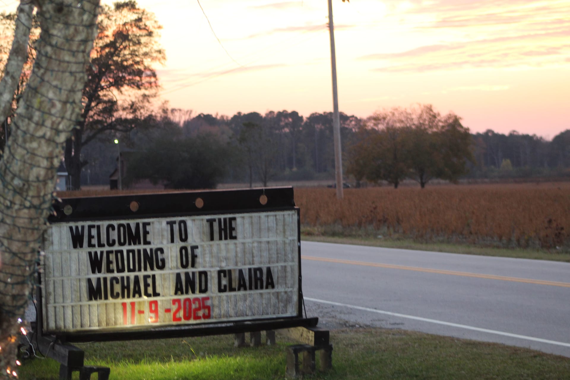 Roadside marquee sign reading Welcome to the Wedding of Michael and Claira 11-9-2025 at dusk with golden farmland fields and a pink sunset sky in the background.