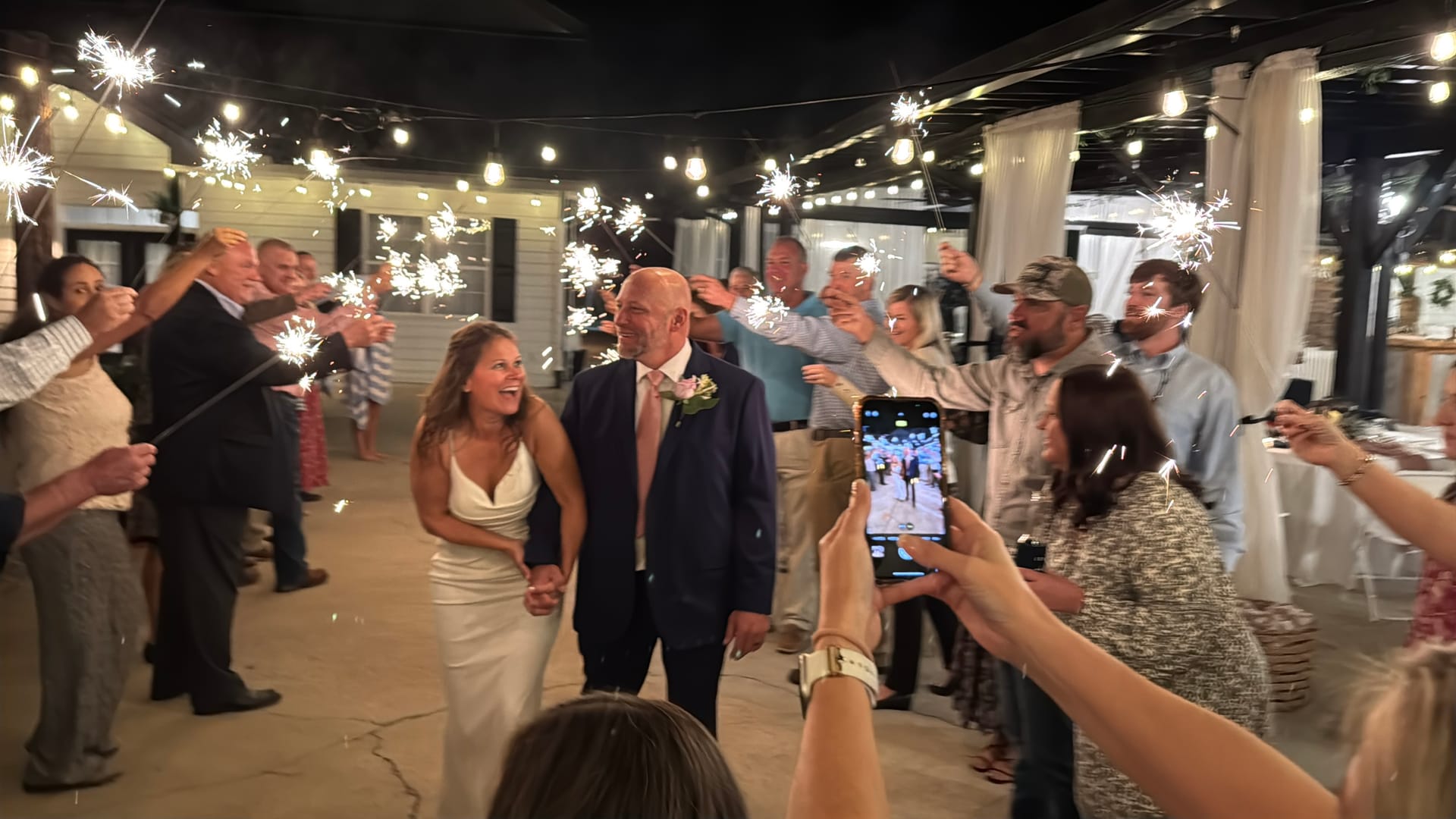 Bride and groom walk hand-in-hand through a glowing sparkler tunnel lined with cheering guests under warm string lights at Scarborough House wedding venue.