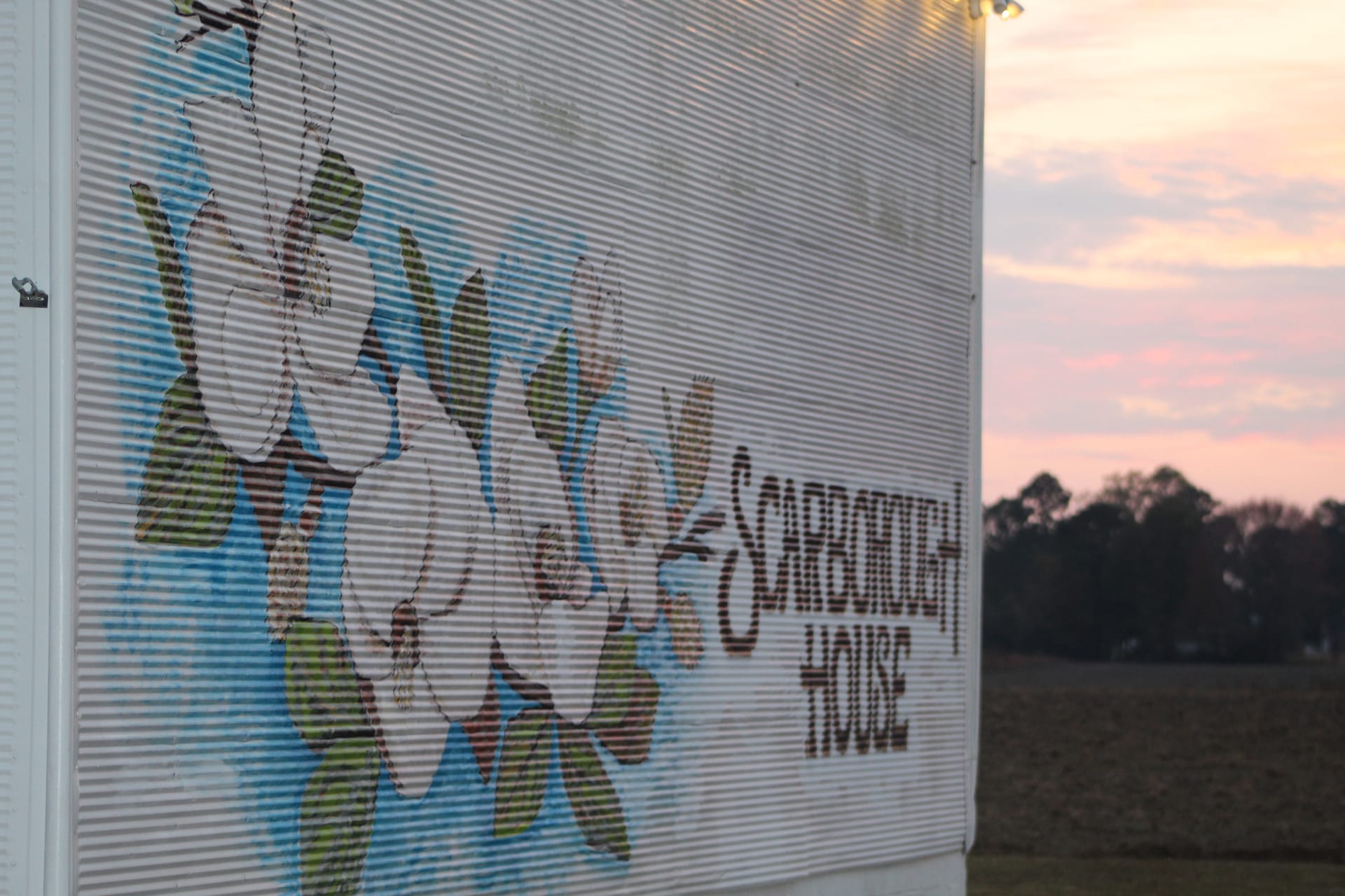Scarborough House painted mural and signage on a corrugated metal silo glowing at sunset with a colorful pink and orange sky and treeline in the background.