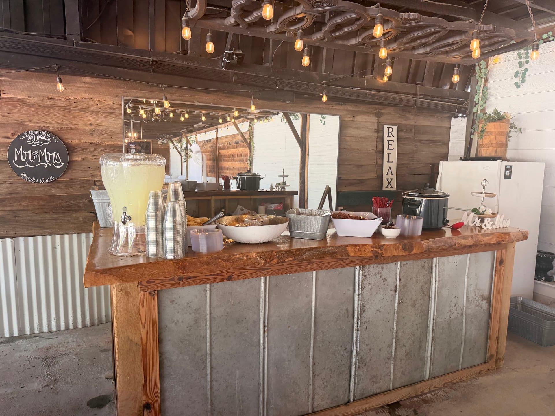 Rustic wedding bar with a live-edge wood countertop, corrugated metal front, lemonade dispenser, buffet spread, and warm Edison bulb string lights at a farmhouse-style wedding venue.