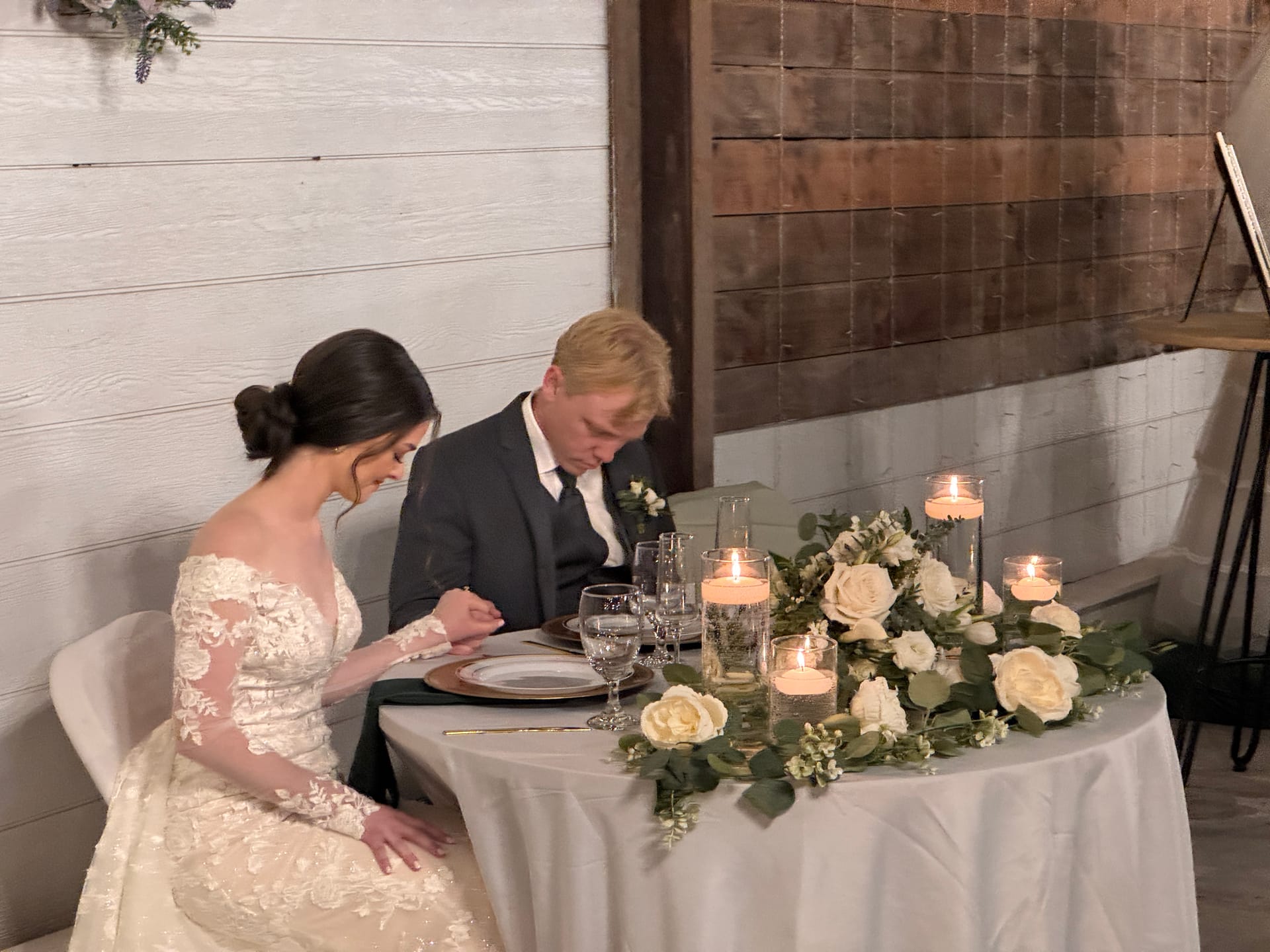 Bride and groom bow their heads in prayer at a candlelit sweetheart table adorned with white roses and eucalyptus garland inside the Scarborough House grand hall.