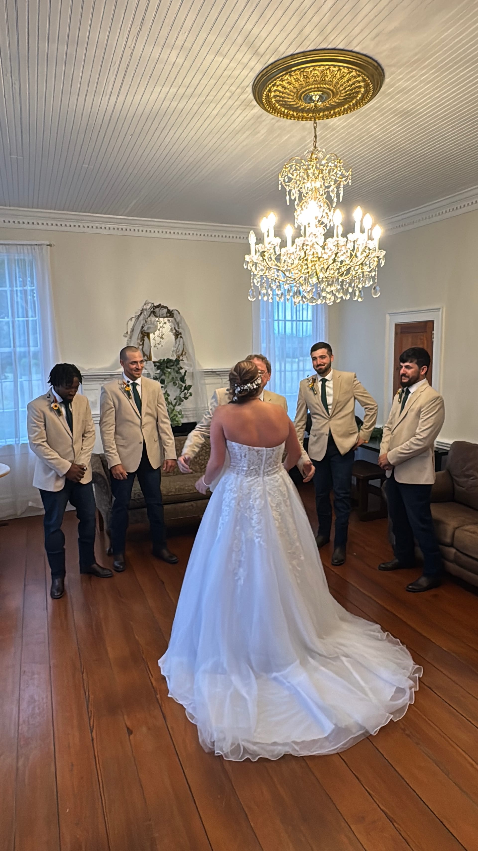 Bride in a white strapless gown stands with her back to the camera as four smiling groomsmen in tan blazers react to her reveal inside the historic grand hall of Scarborough House beneath a crystal chandelier.