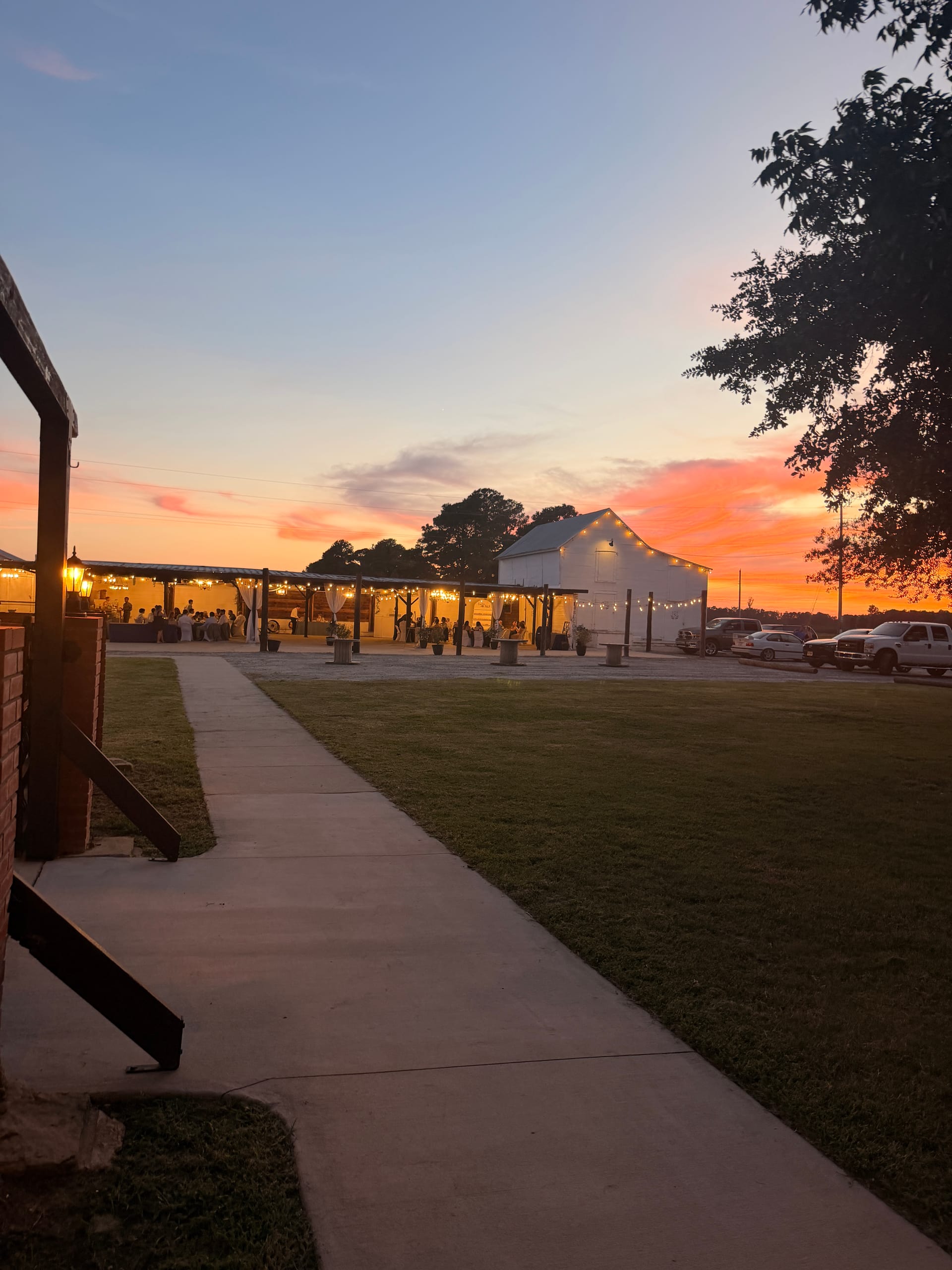 Warm sunset sky glowing orange and pink behind a white barn wedding venue adorned with string lights and guests gathered under a rustic open-air pavilion at Scarborough House.