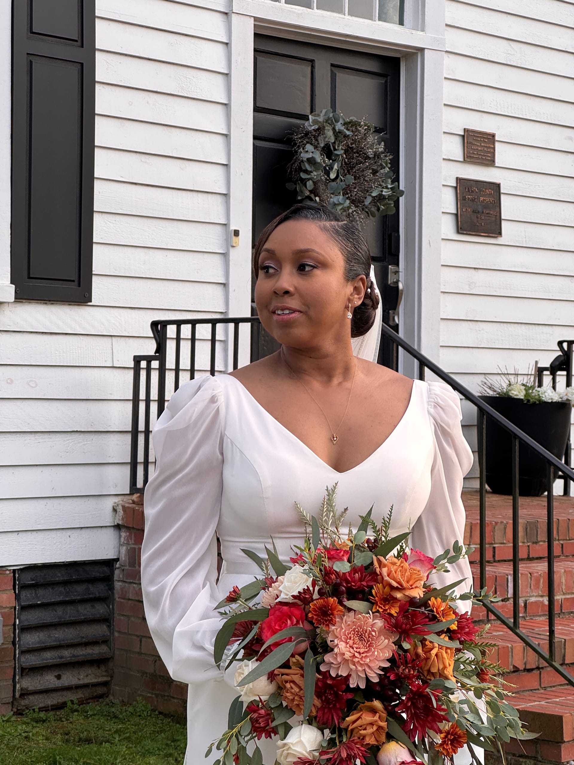 Bride in a white long-sleeve gown holding a cascading autumn bouquet of deep red, orange, and blush blooms stands before the historic white clapboard facade of Scarborough House.