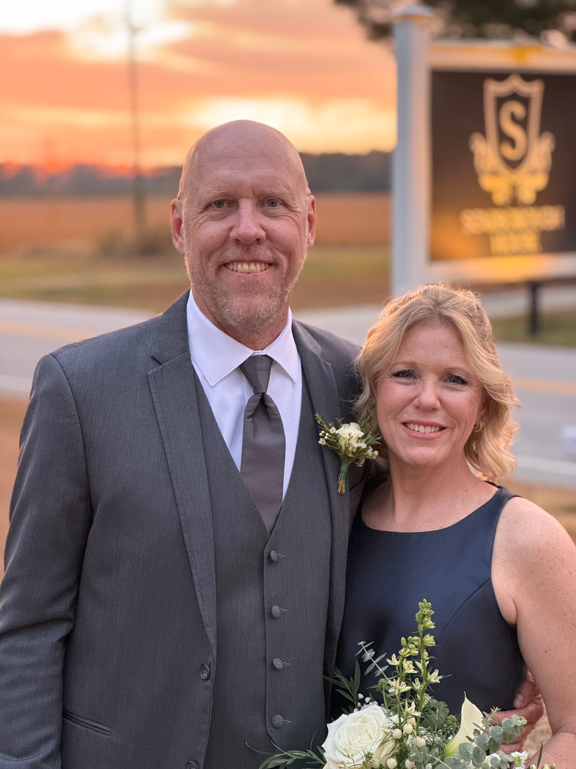 Elegantly dressed couple smiling together in front of the Scarborough House venue sign at golden-hour sunset, man in grey suit with boutonniere and woman in navy dress holding white floral bouquet.
