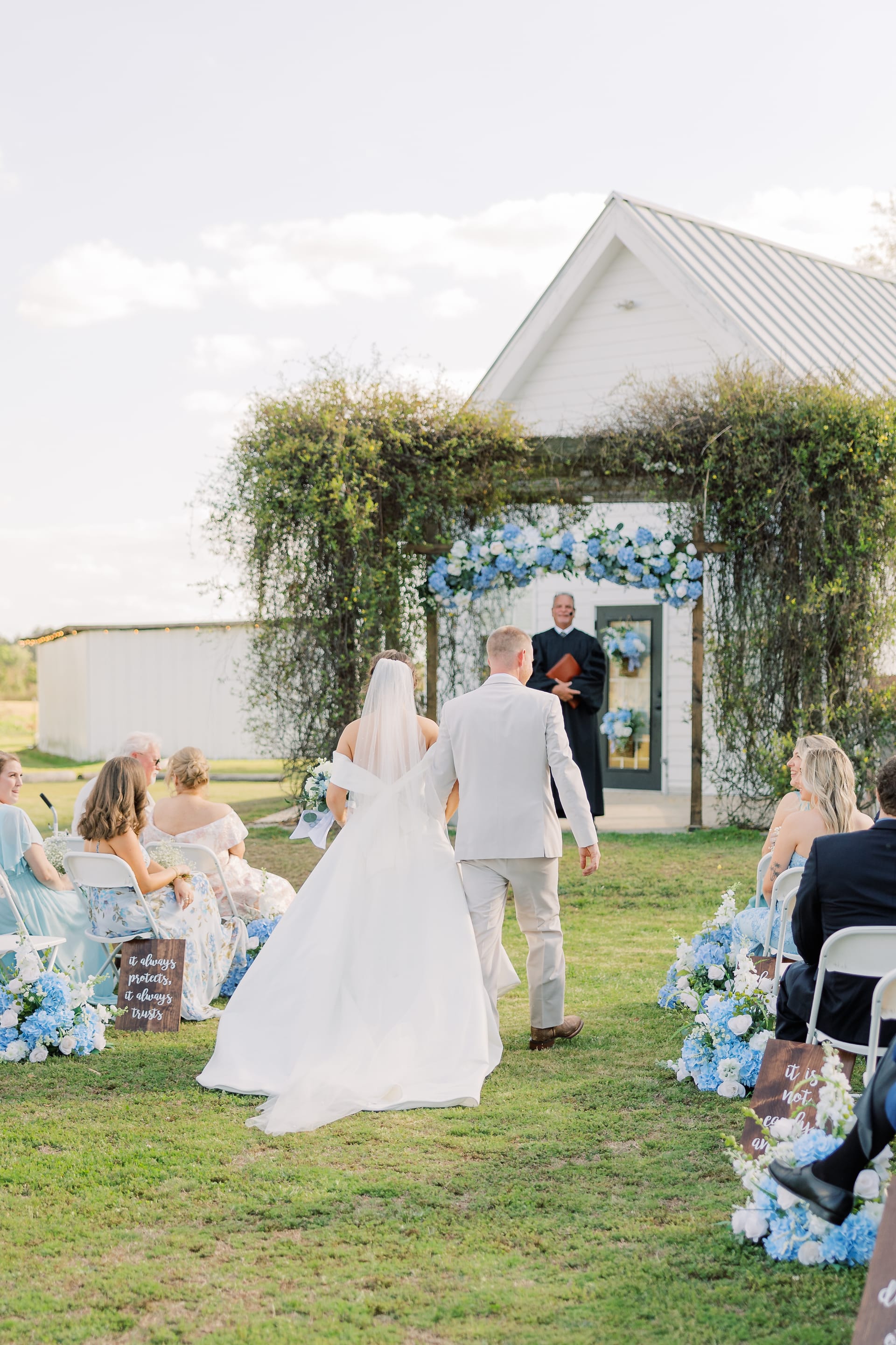 Bride and groom walk down the outdoor aisle toward a smiling officiant beneath a vine-covered white chapel adorned with blue hydrangea floral arrangements at Scarborough House.