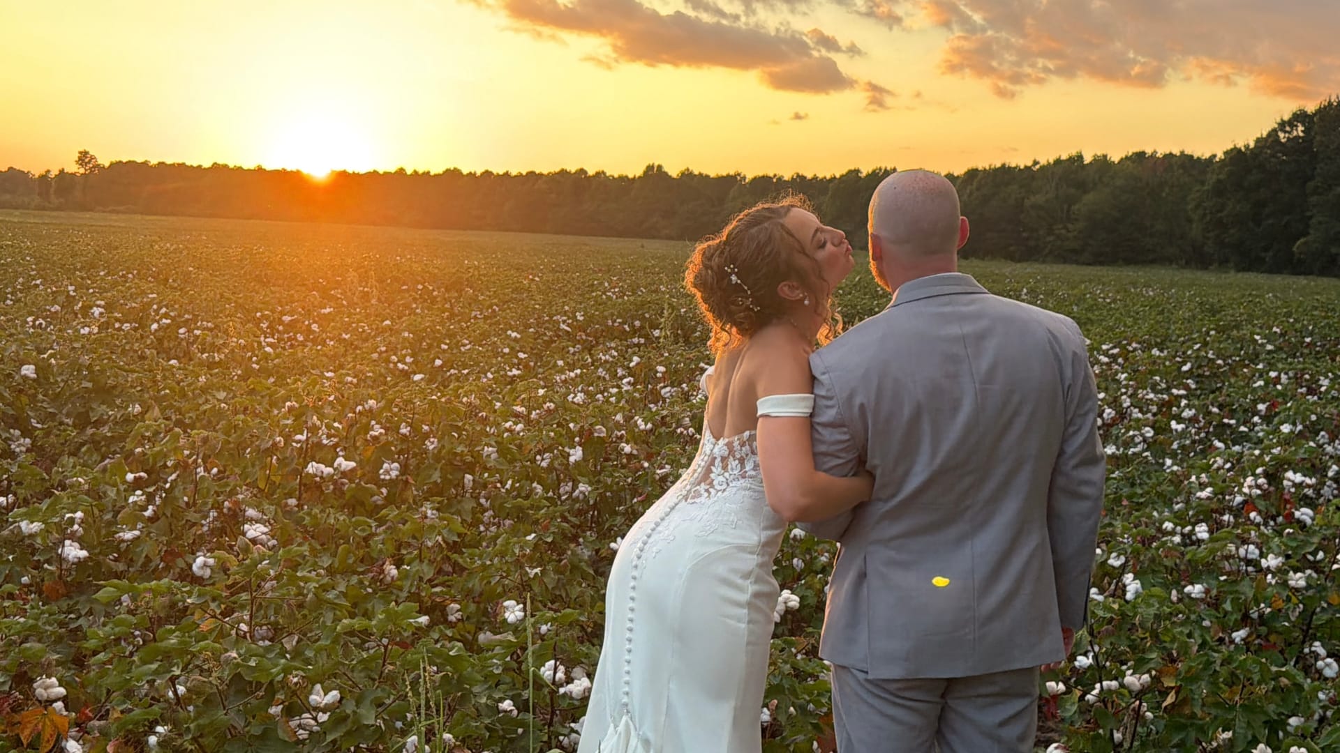 Bride in a lace off-shoulder gown and groom in a gray suit embrace in a blooming cotton field at Scarborough House as a vivid golden sunset glows on the horizon.