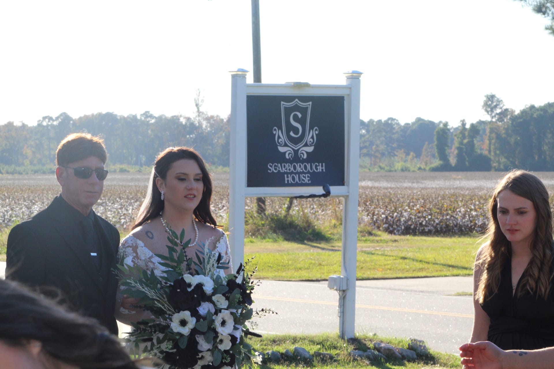 Bride holding black and white floral bouquet stands beside the Scarborough House marquee sign with a cotton field backdrop on her wedding day.