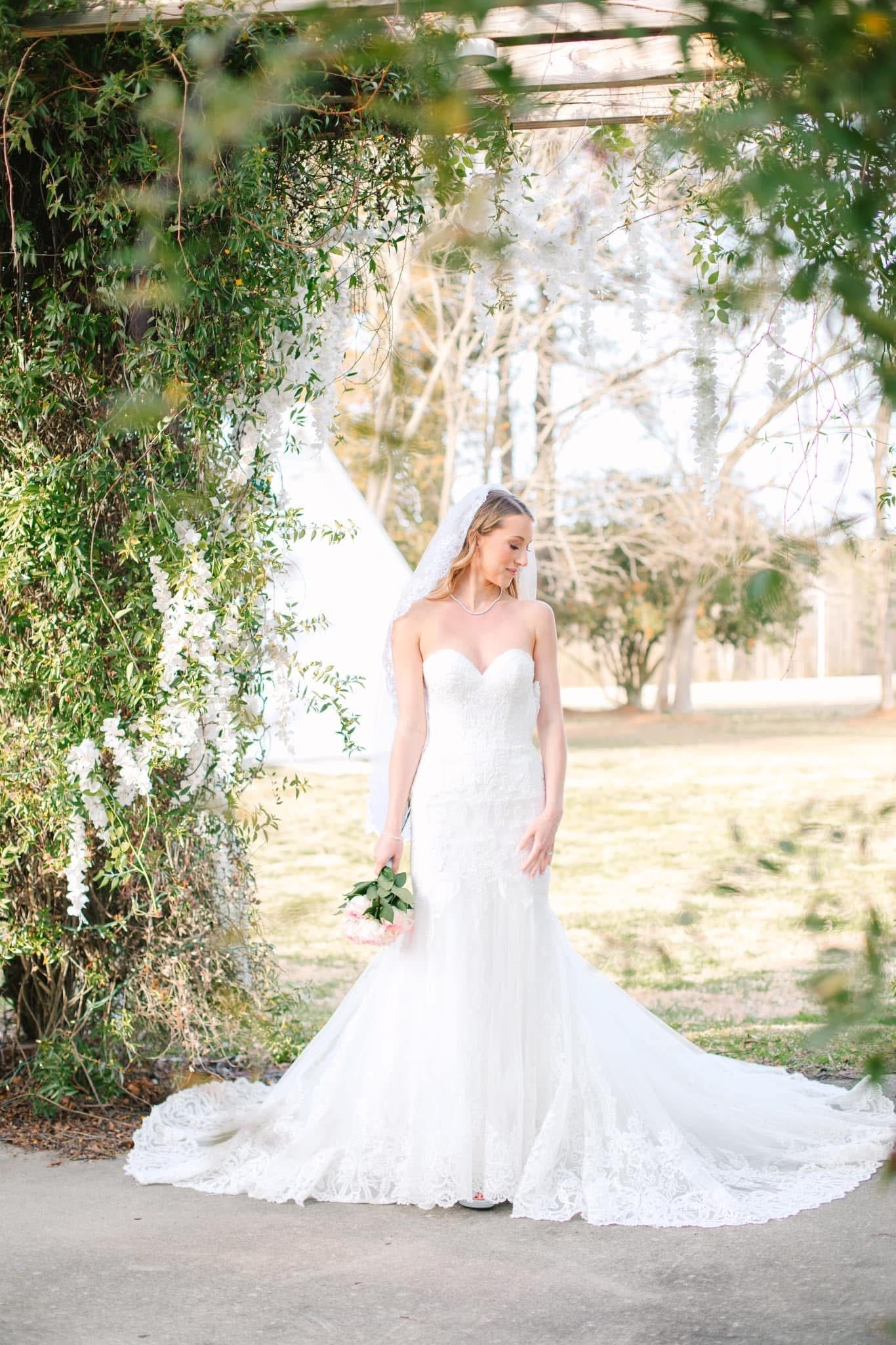 Bride in a strapless lace mermaid gown and veil holding a pink bouquet stands beneath a floral and greenery archway at Scarborough House historic estate.