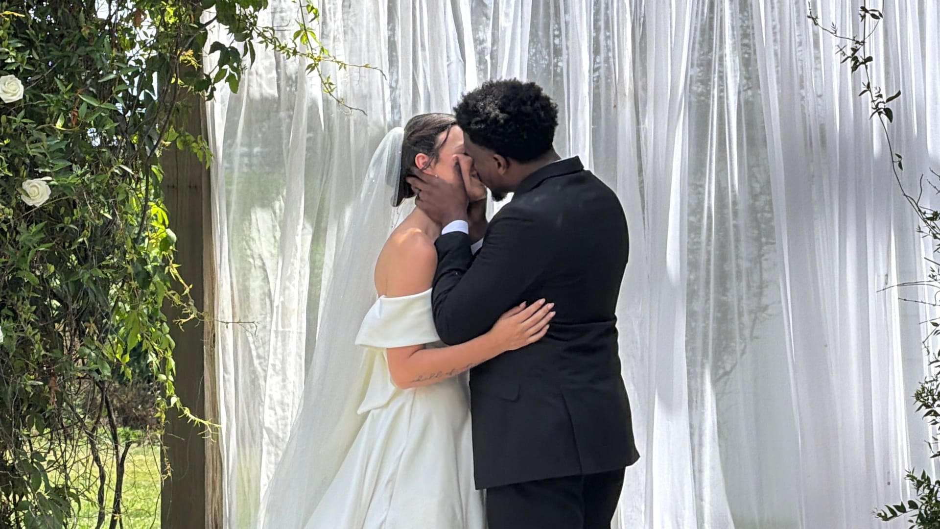 Bride in off-shoulder white gown and groom in black suit share their first kiss beneath flowing white drape backdrop at Scarborough House wedding ceremony.