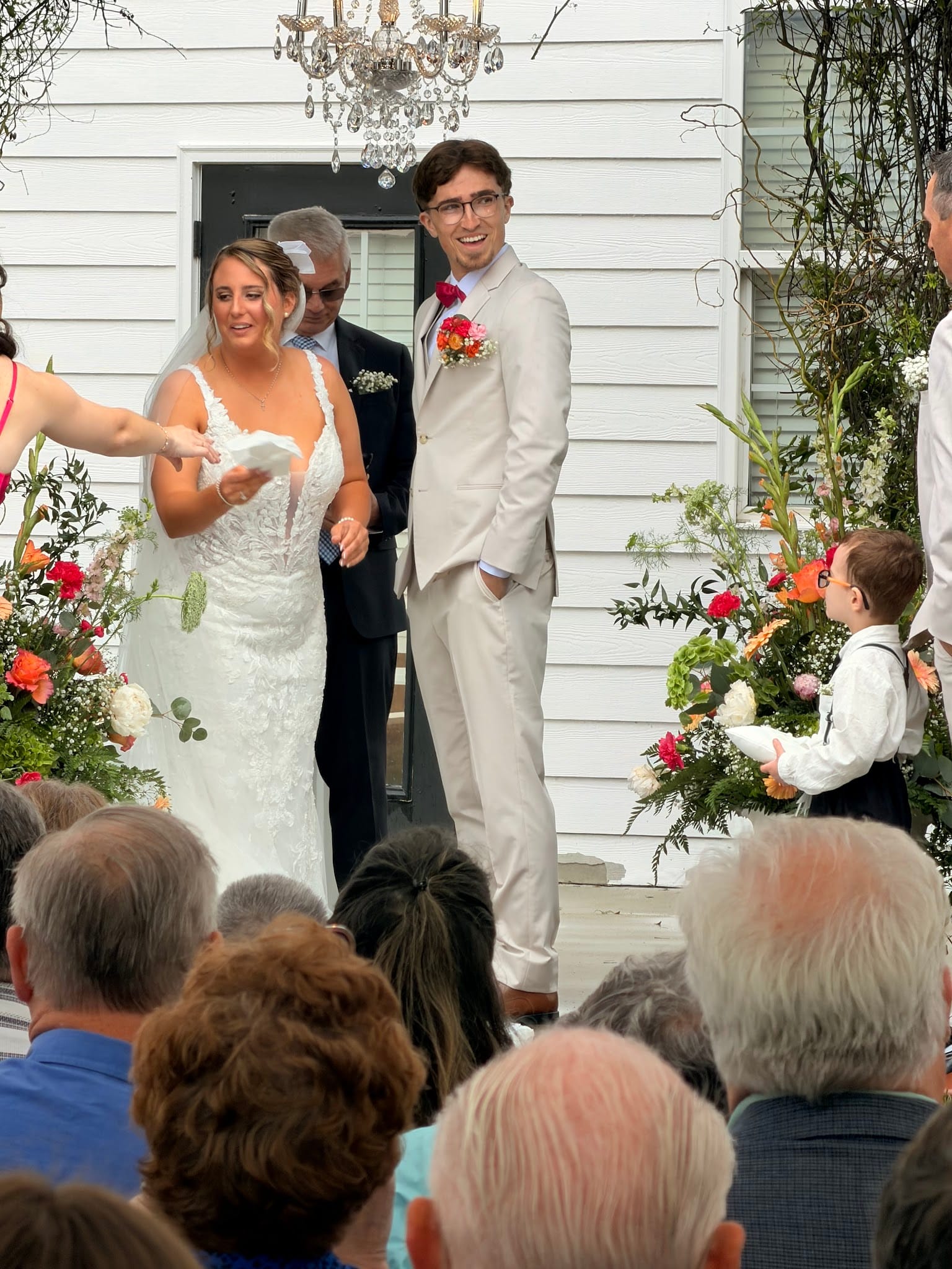 Smiling bride in lace gown and groom in tan suit descend the front steps of the historic 1821 Scarborough House manor surrounded by vibrant floral arrangements during their wedding recessional.